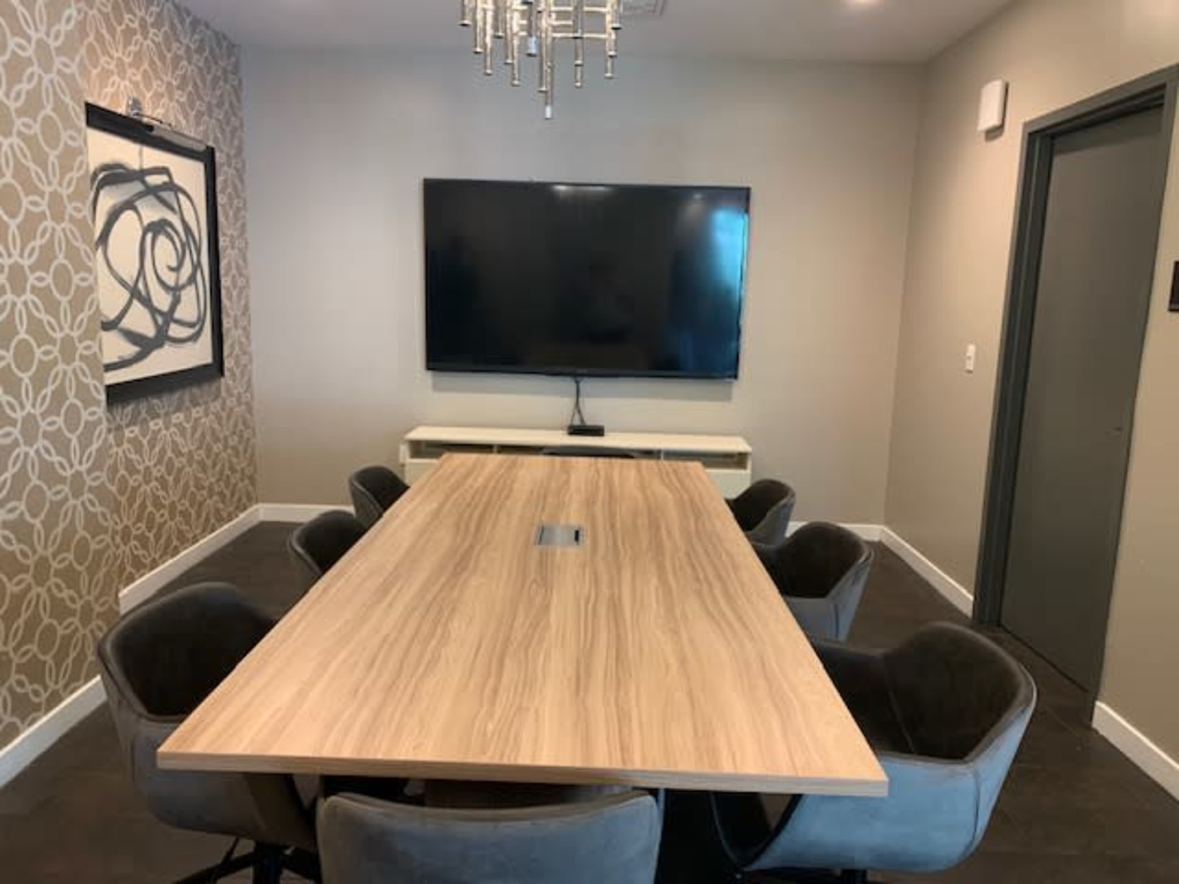 A modern conference room features a large wooden table surrounded by upholstered chairs, with a wall-mounted television and decorative wallpaper.