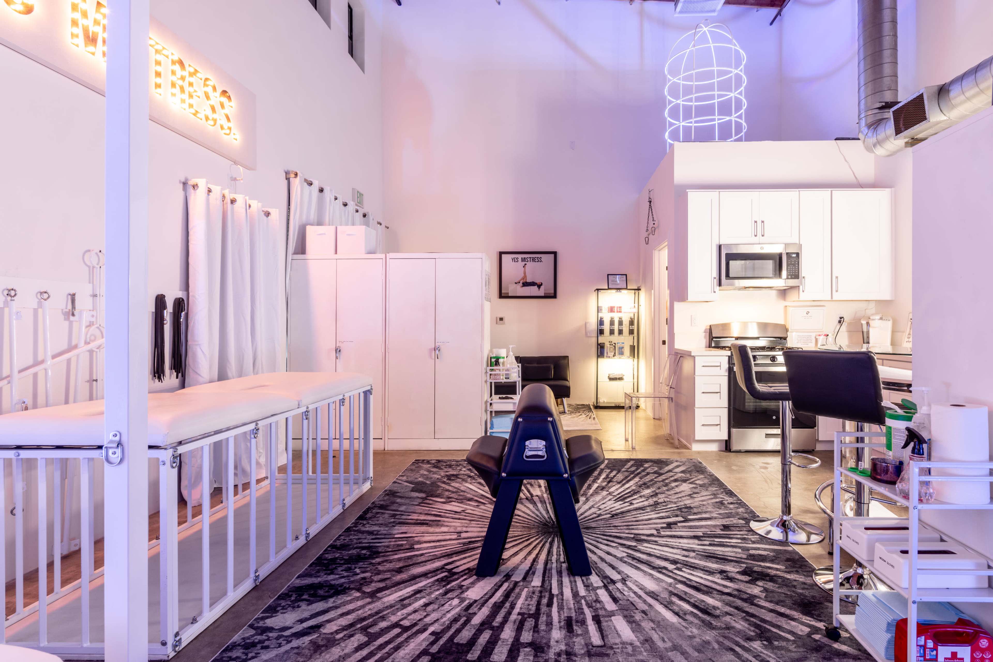 The image shows a modern treatment space with a massage table, a kitchenette, and various clinical equipment organized throughout the room.