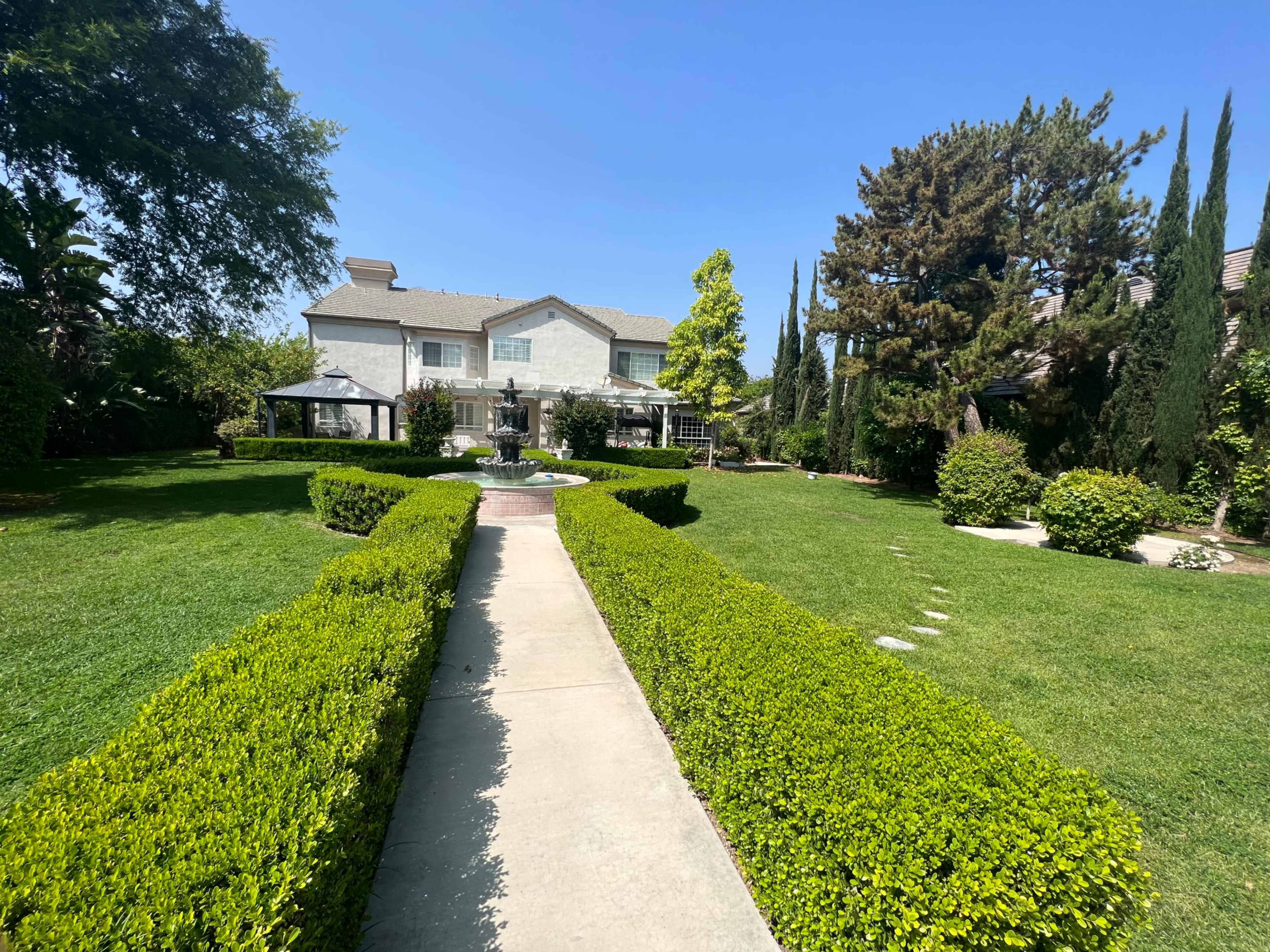 A neatly landscaped pathway lined with hedges leads to a large building surrounded by trees and decorative features.