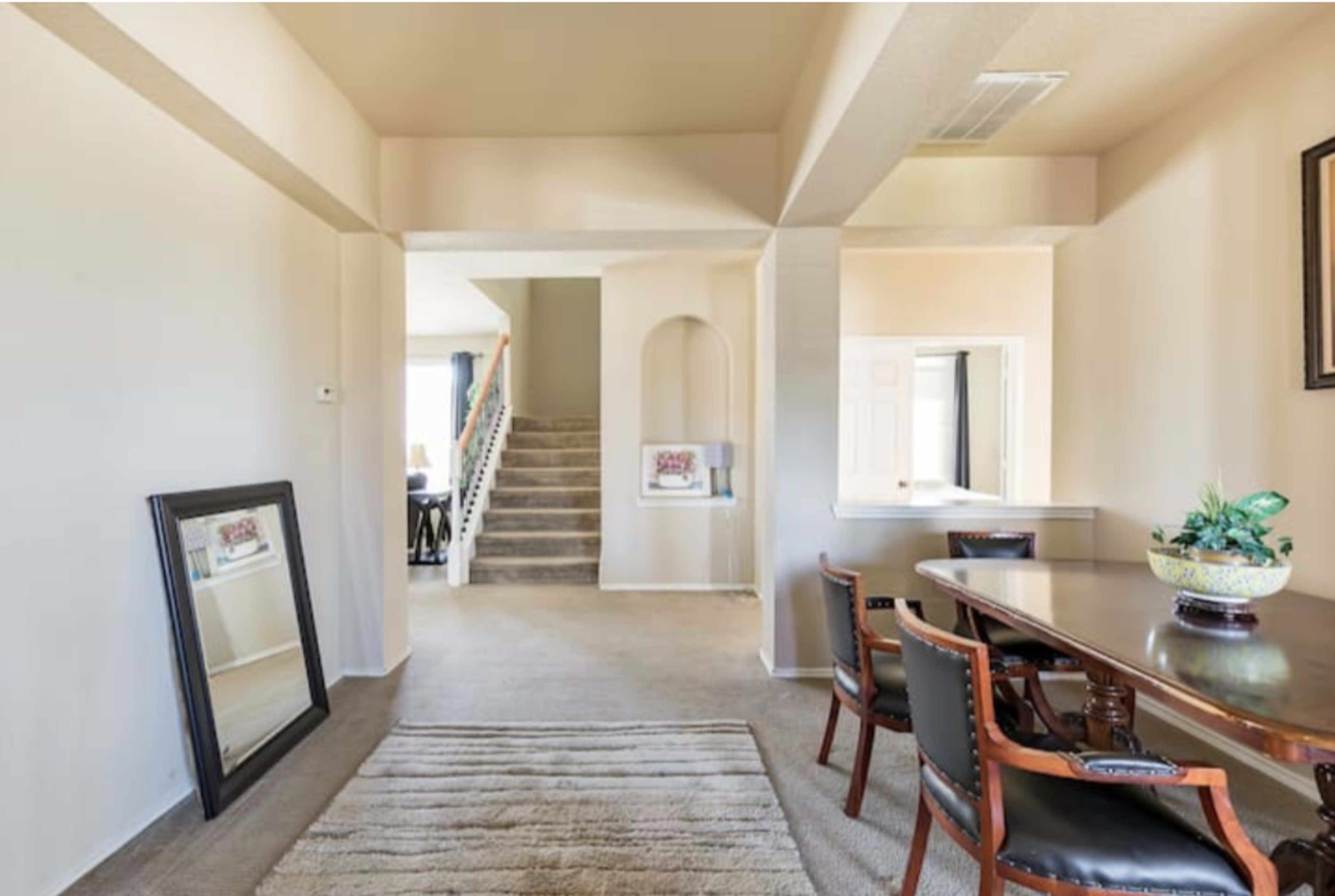 A hallway with a staircase, a dining table with chairs, a large mirror leaning against the wall, and light-colored walls.