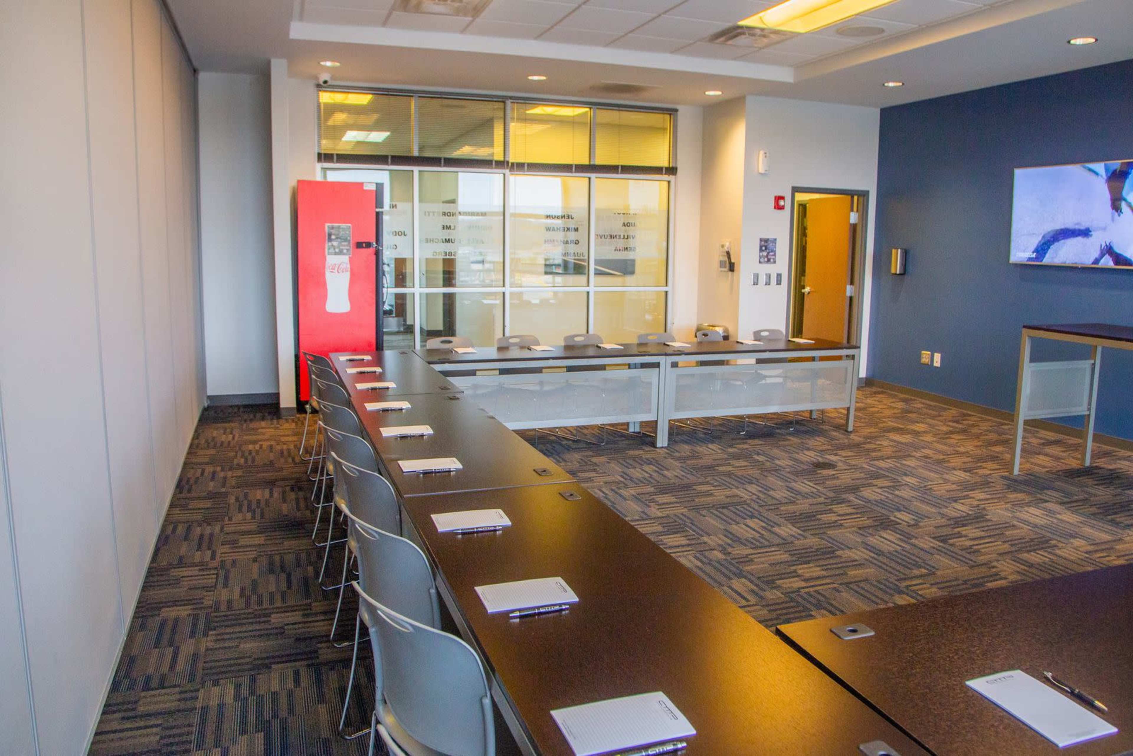 A conference room features a long table with chairs, a vending machine, and a wall-mounted screen.