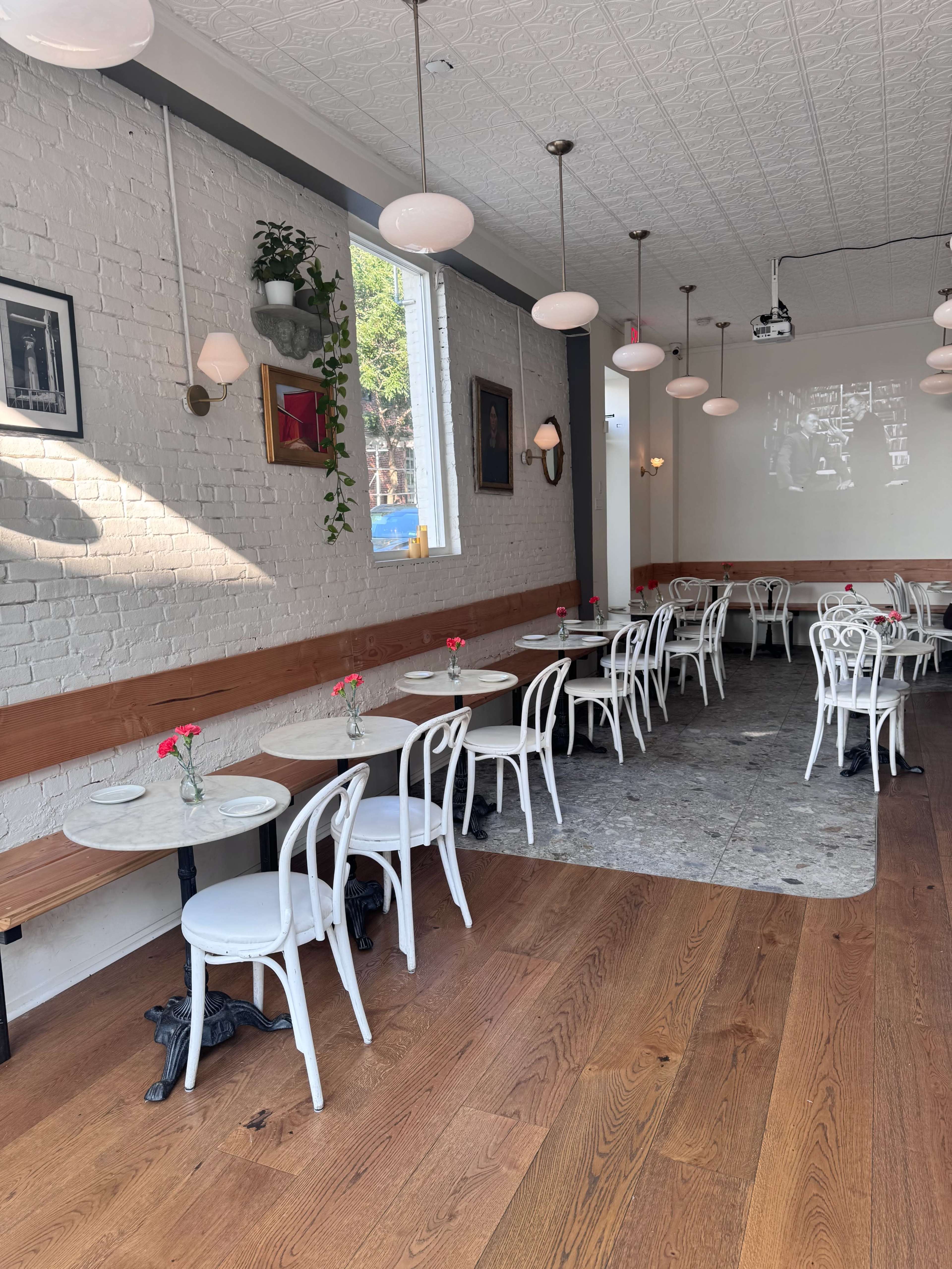 The image shows a bright, modern café interior with white walls, wooden flooring, and small tables set for patrons, each adorned with a single flower in a vase.