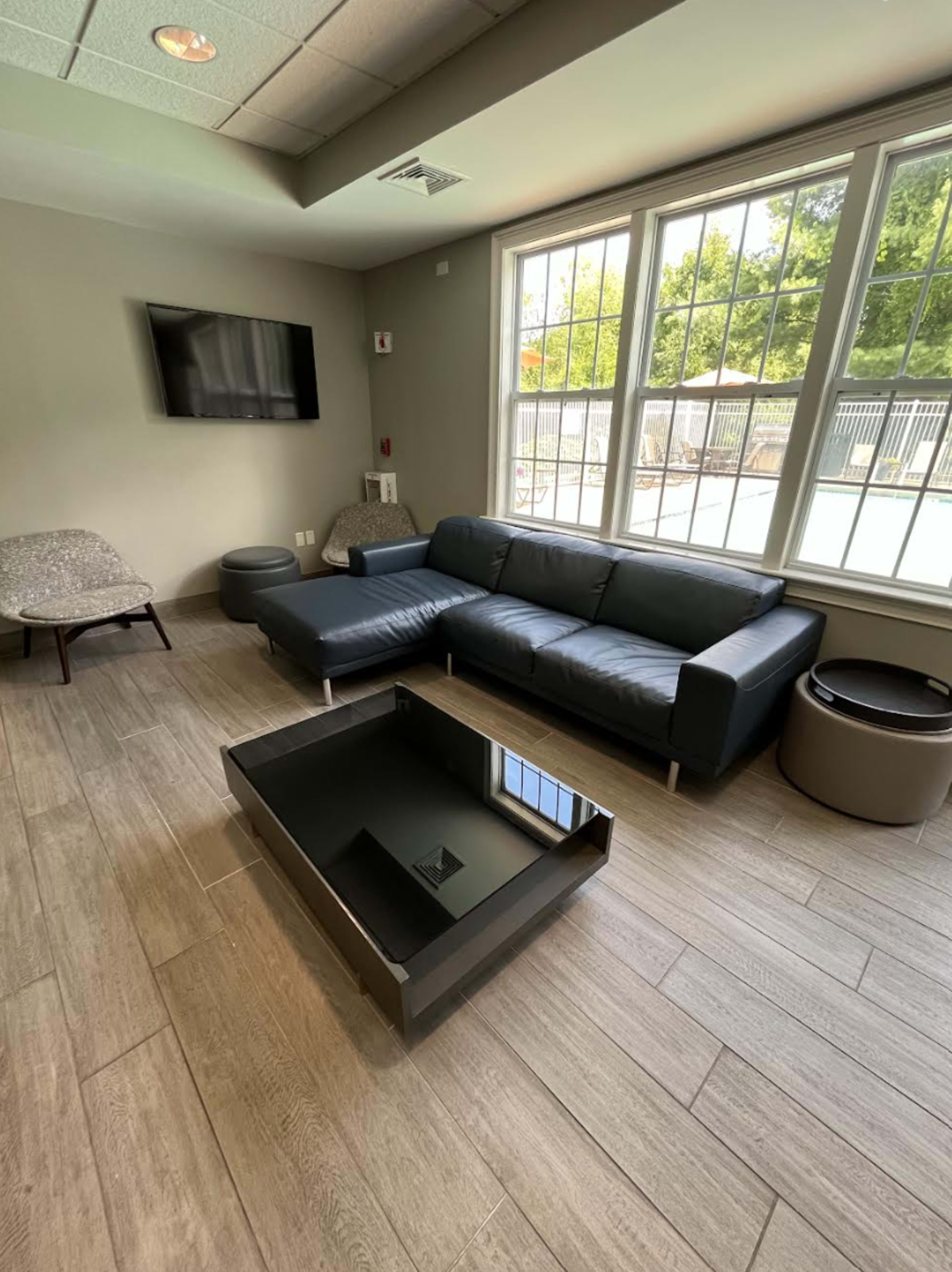 The image shows a modern sitting area with a black leather sofa, a glass-top coffee table, and a large window overlooking an outdoor space.