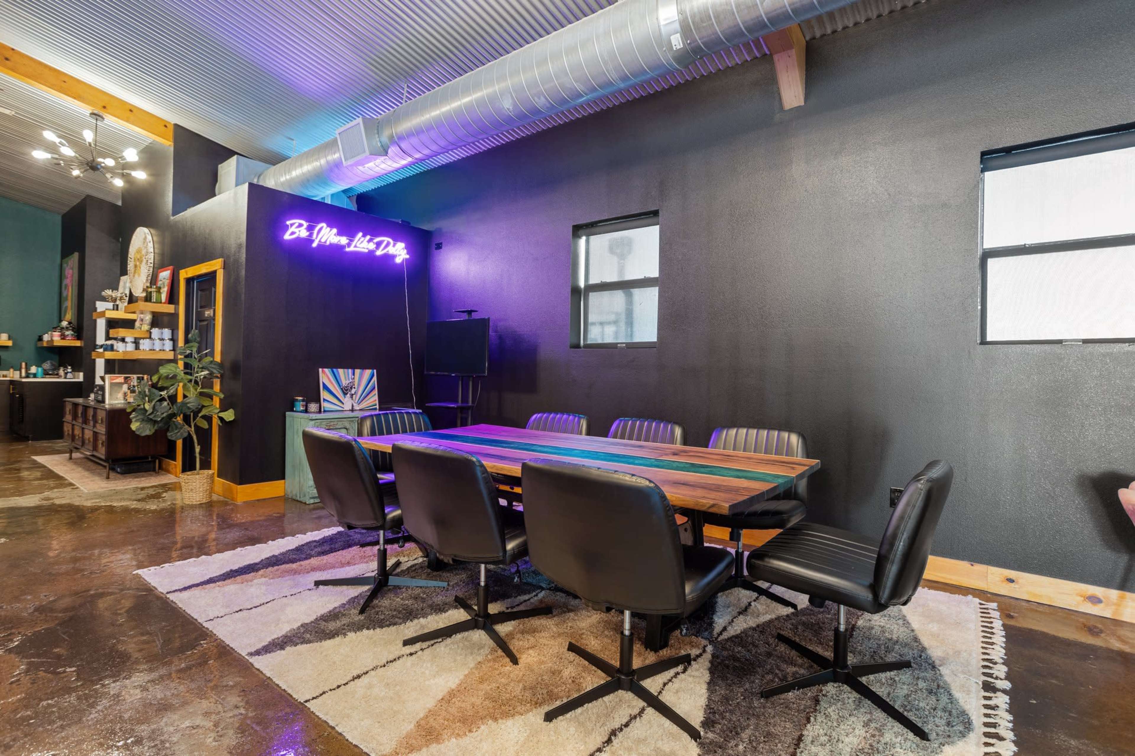 A modern meeting room featuring a long wooden table surrounded by black chairs, with a neon sign on the wall and large windows letting in natural light.