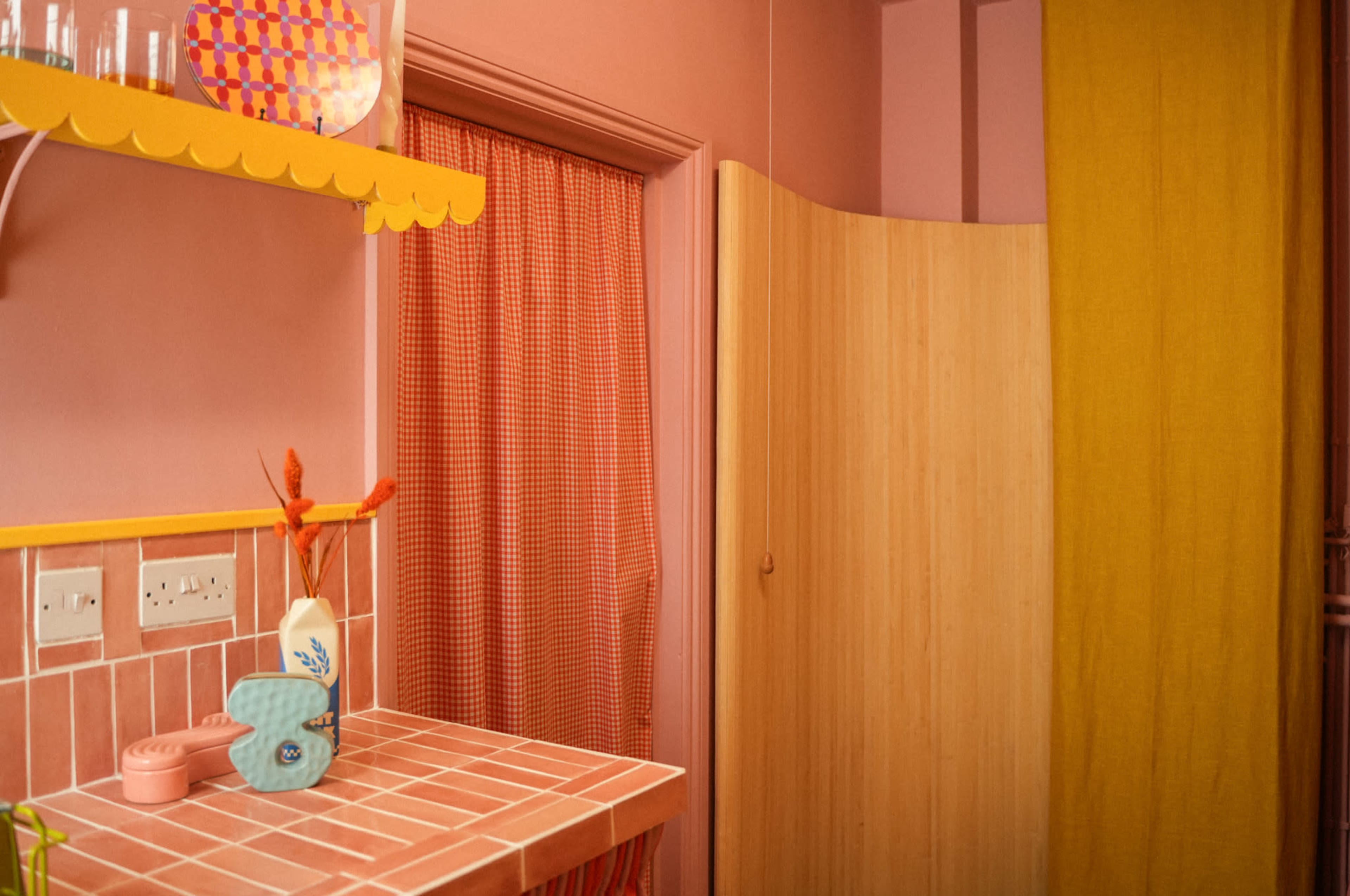 The image depicts a pastel-colored kitchen space with a tiled counter, whimsical dishware, and a yellow curtain partially covering a doorway.