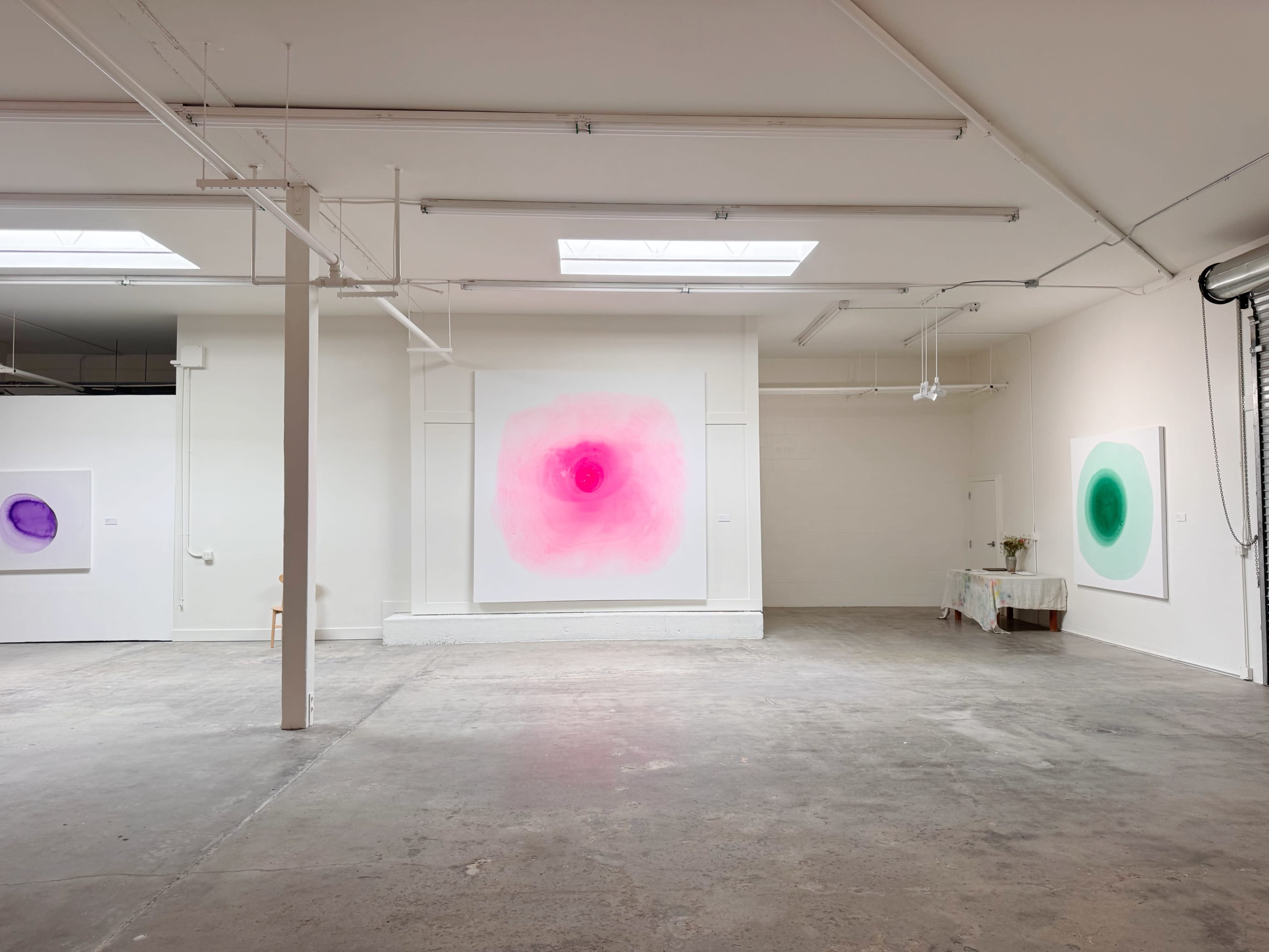 The image shows an art gallery with two large, abstract paintings hanging on white walls and a minimalistic space featuring a concrete floor.