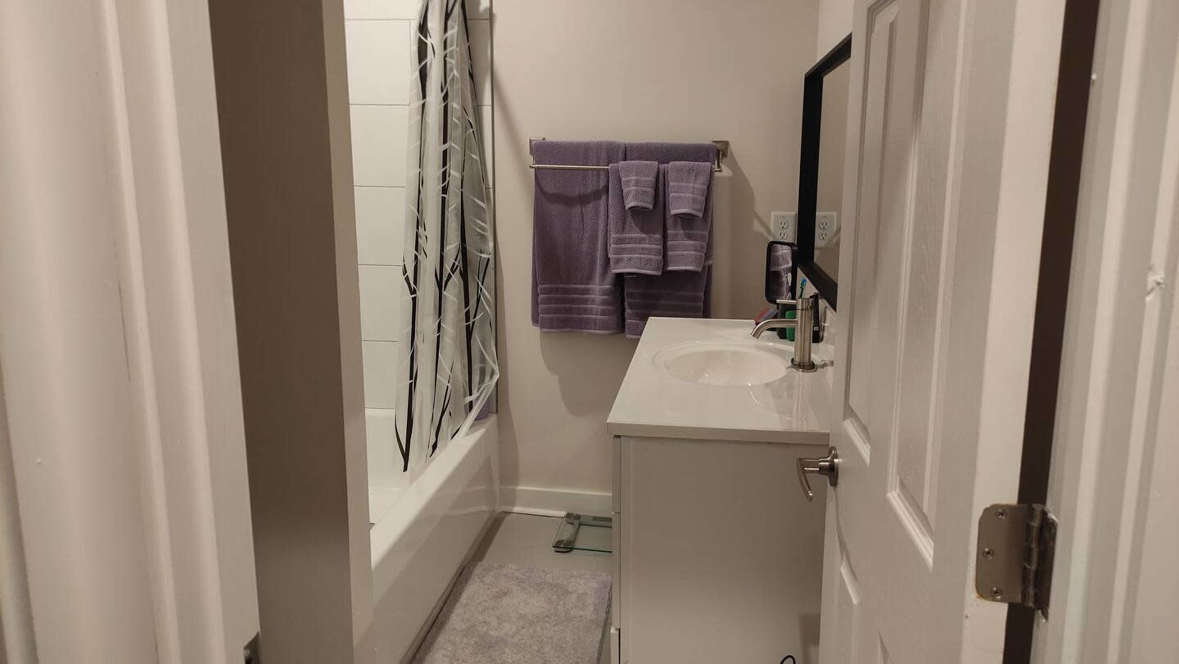The image shows a small bathroom featuring a bathtub, a sink with a mirror, and towels hanging on the wall.