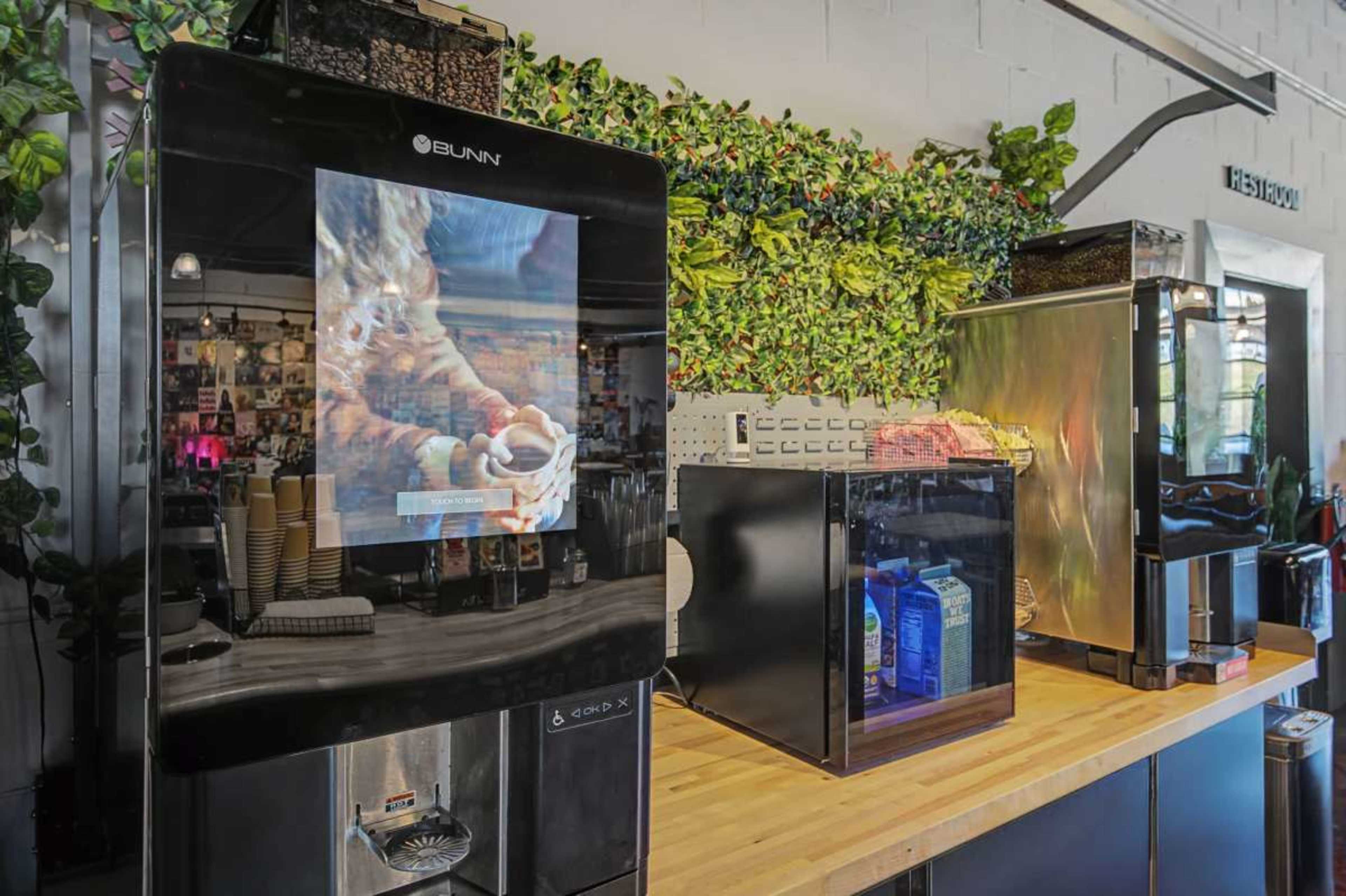 The image shows a coffee station featuring multiple coffee machines on a wooden countertop, with a background of green foliage.