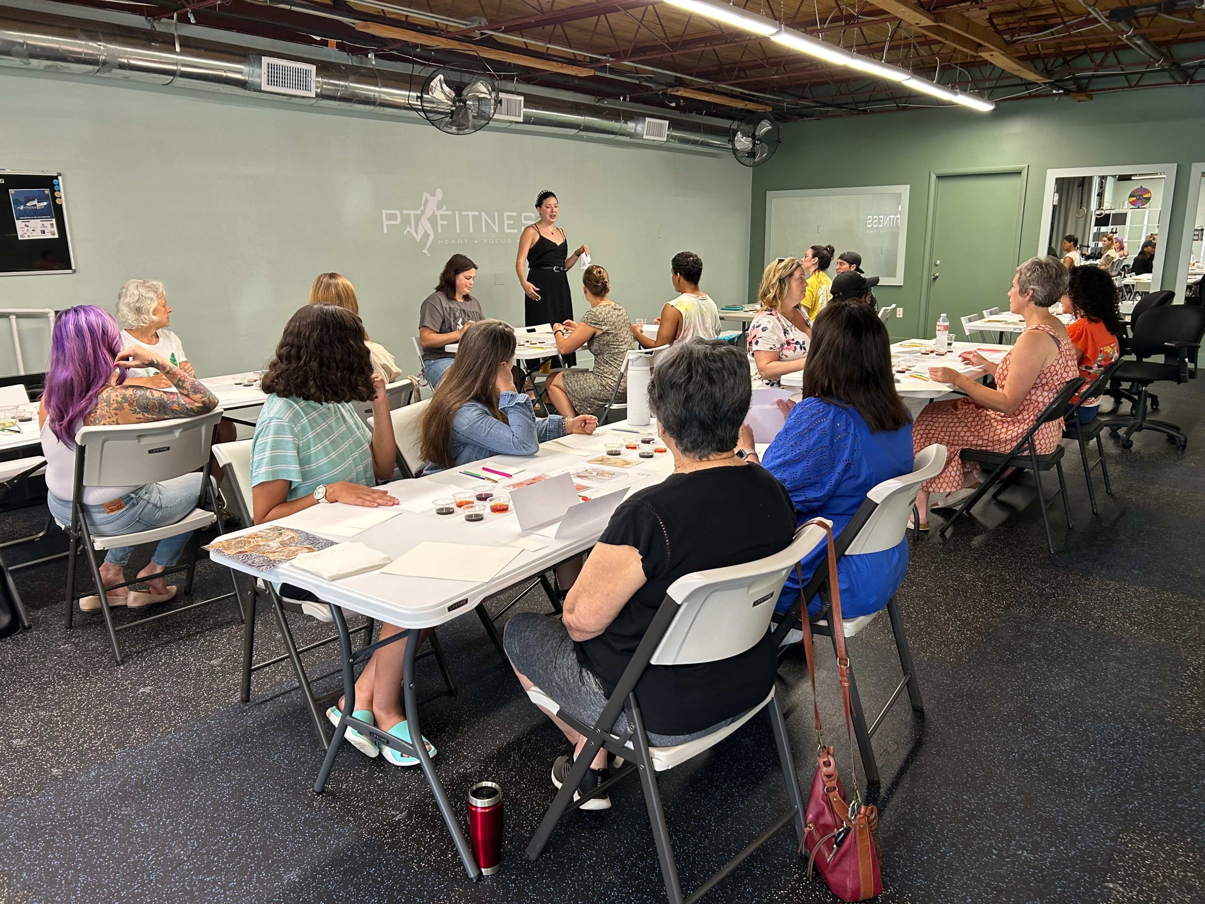A woman stands at the front of a room filled with seated participants engaged in a workshop or class.