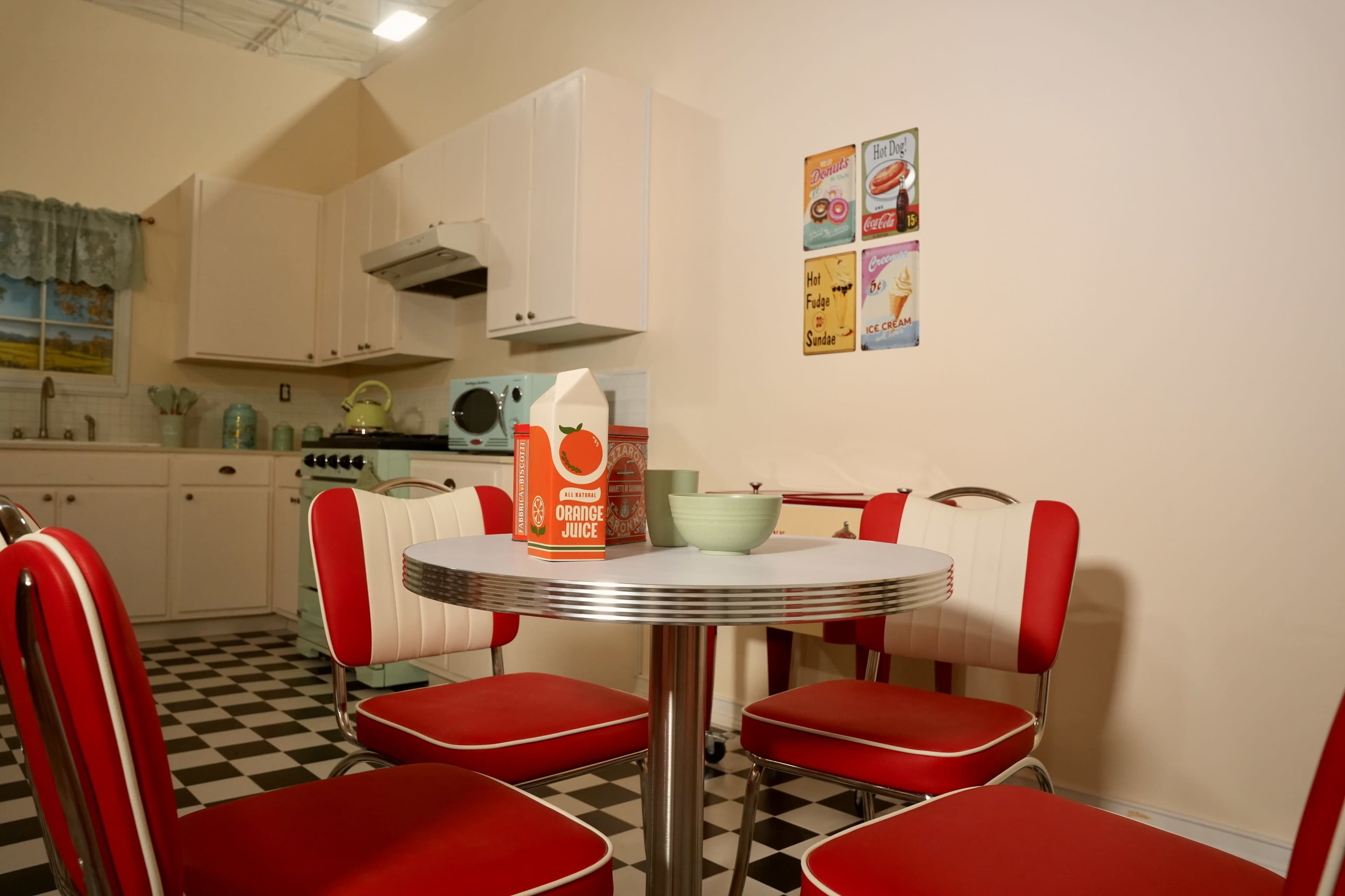 A retro-style kitchen features a round table with red and chrome chairs, surrounded by white cabinets and a checkerboard floor.