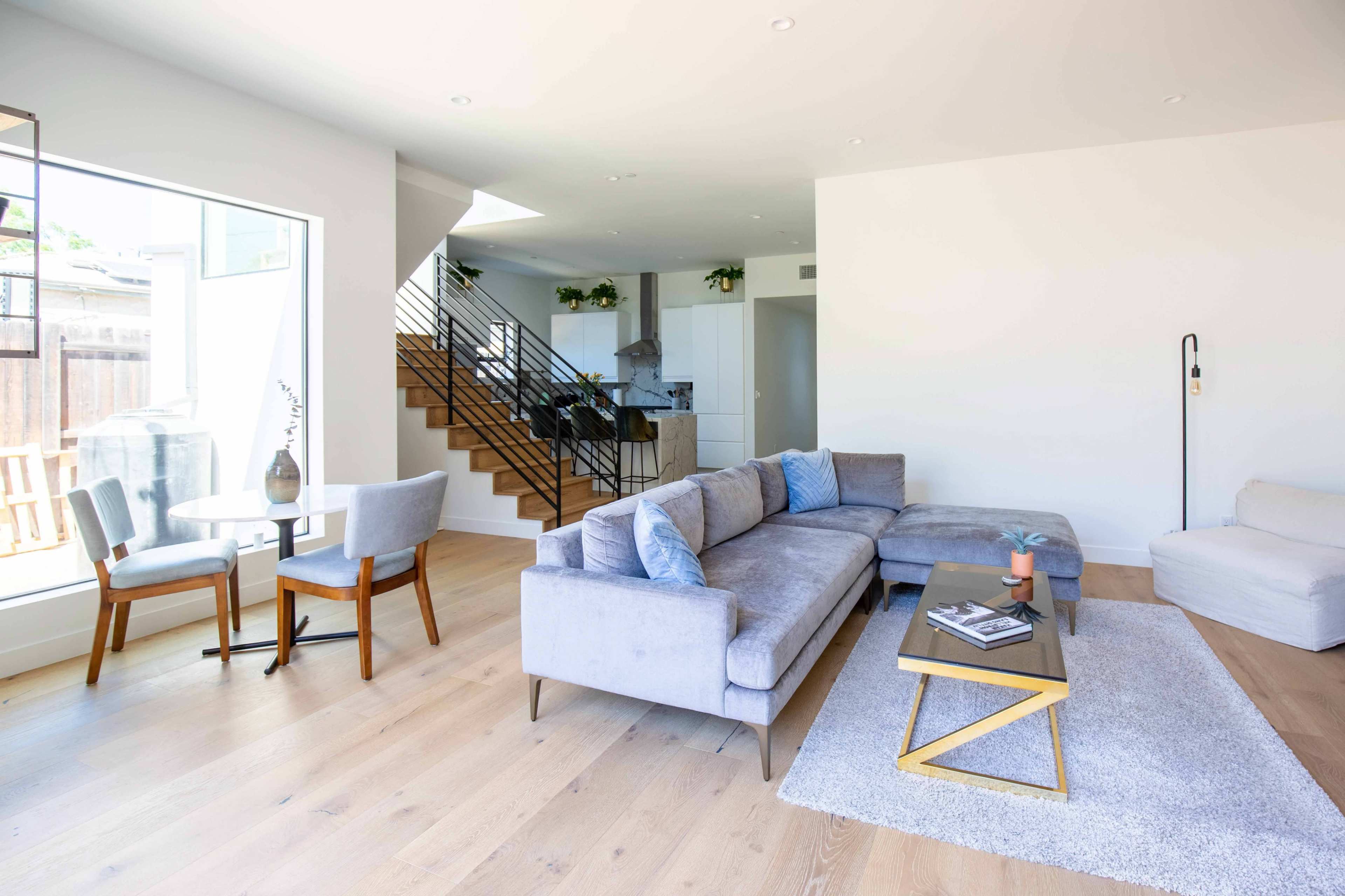 The image depicts a modern living room featuring a gray sectional sofa, a glass coffee table, and a small dining area with two chairs beside a window.