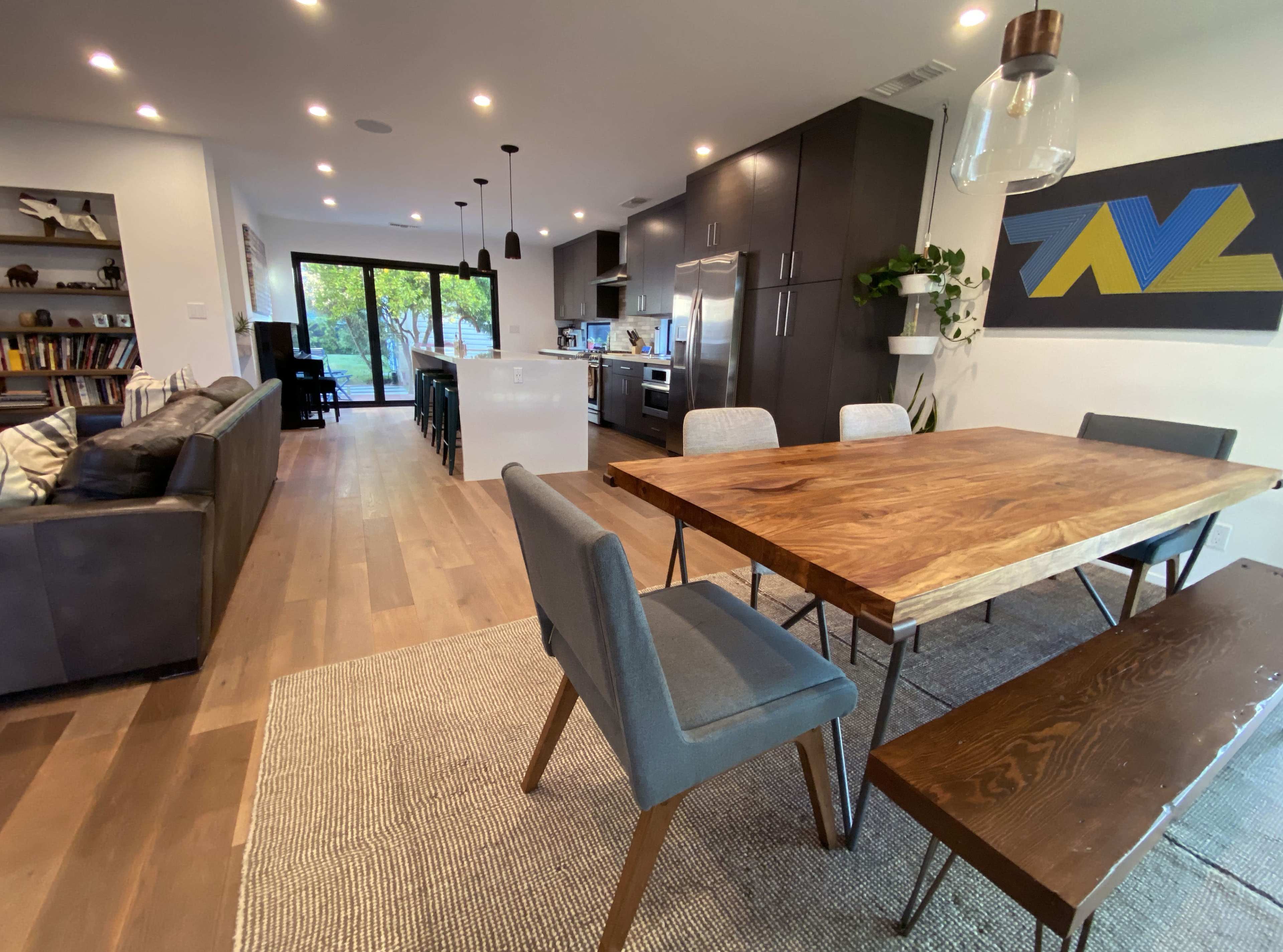 A modern open-concept living and dining area featuring a wooden dining table, gray chairs, and a kitchen in the background with large glass doors leading to an outdoor space.