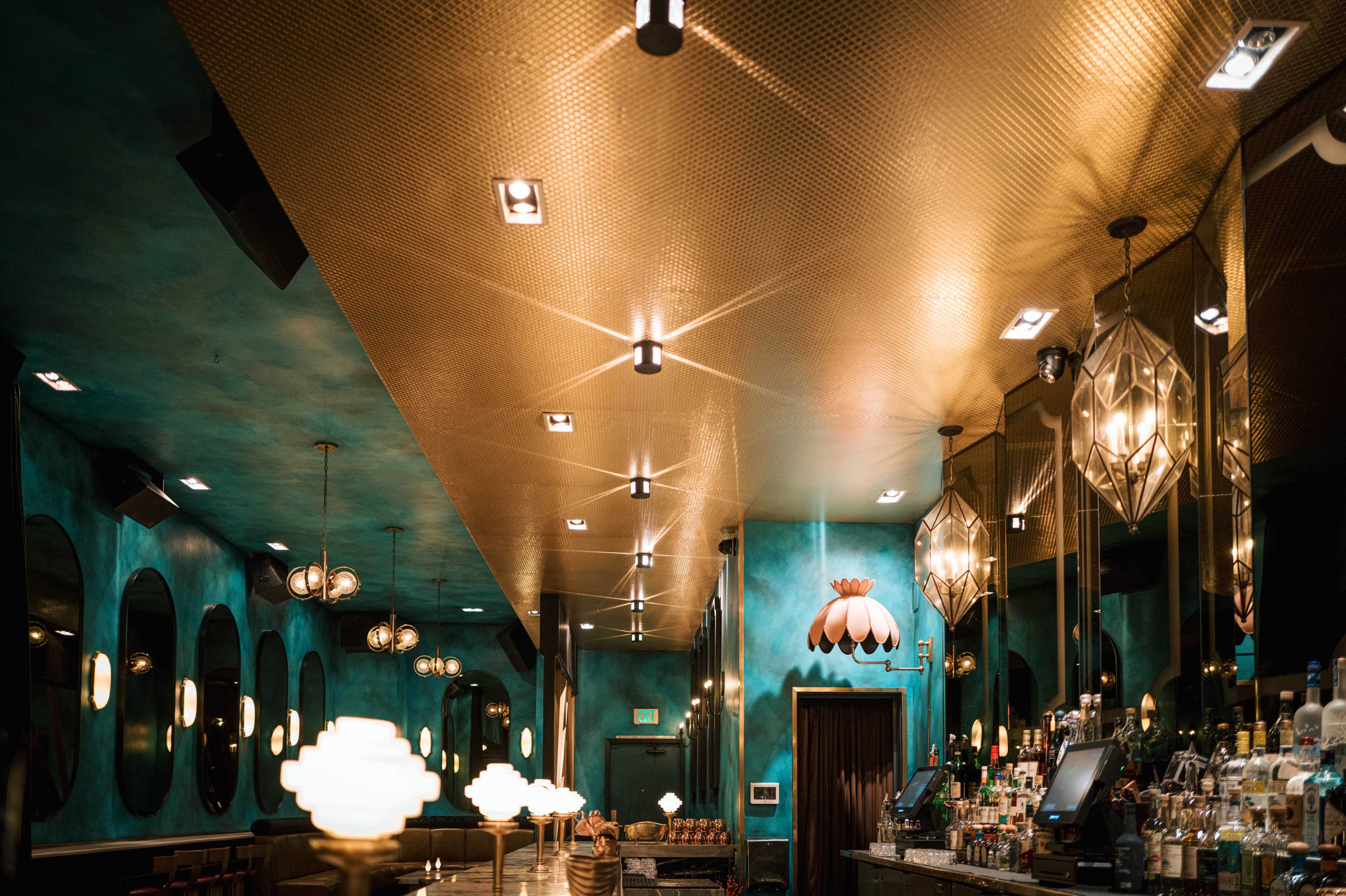 The image shows a bar interior featuring turquoise walls, gold accents on the ceiling, and stylish light fixtures.