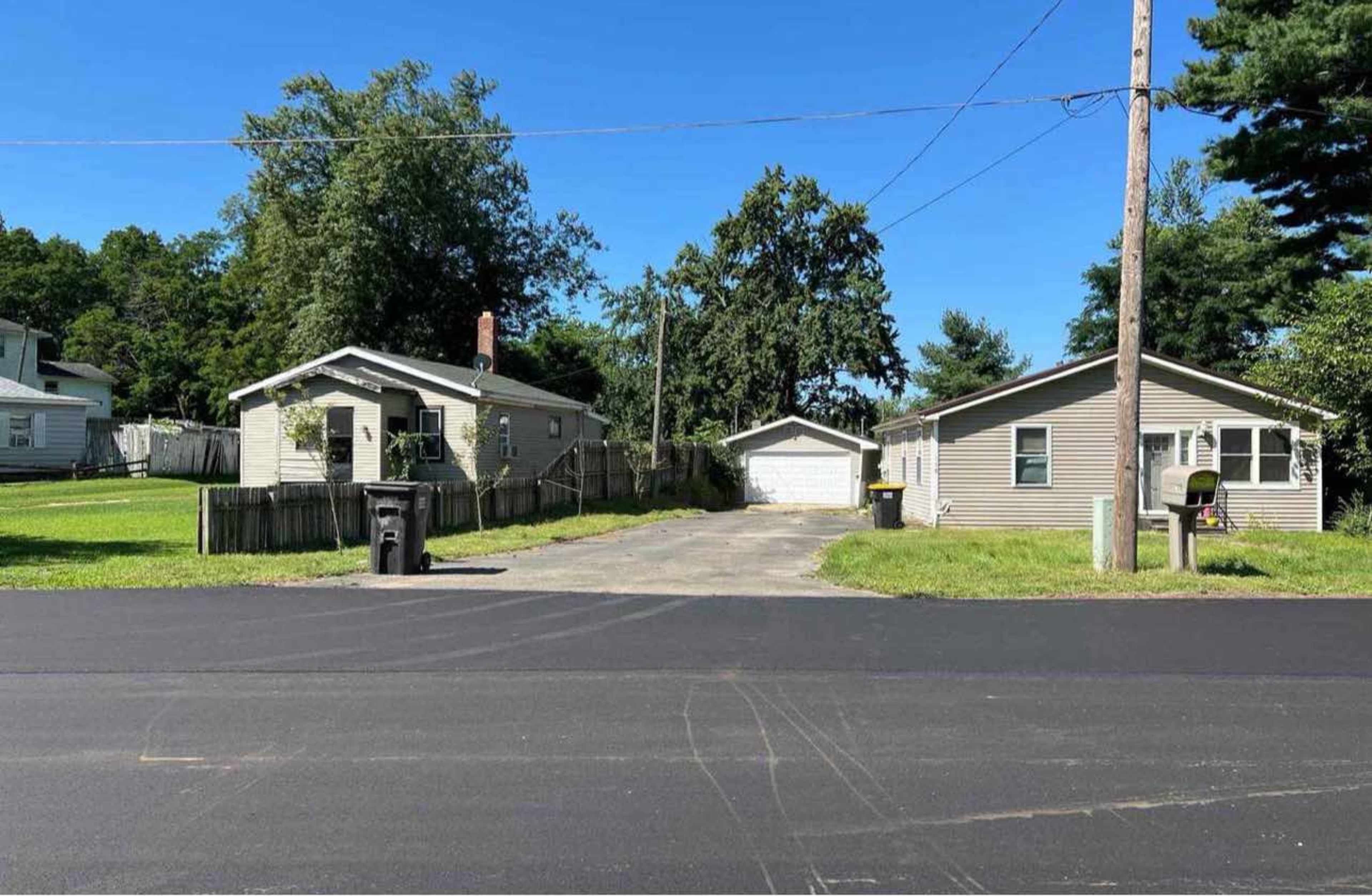 The image shows two houses on opposite sides of a driveway, with a garage in the center, surrounded by trees and a clear blue sky.