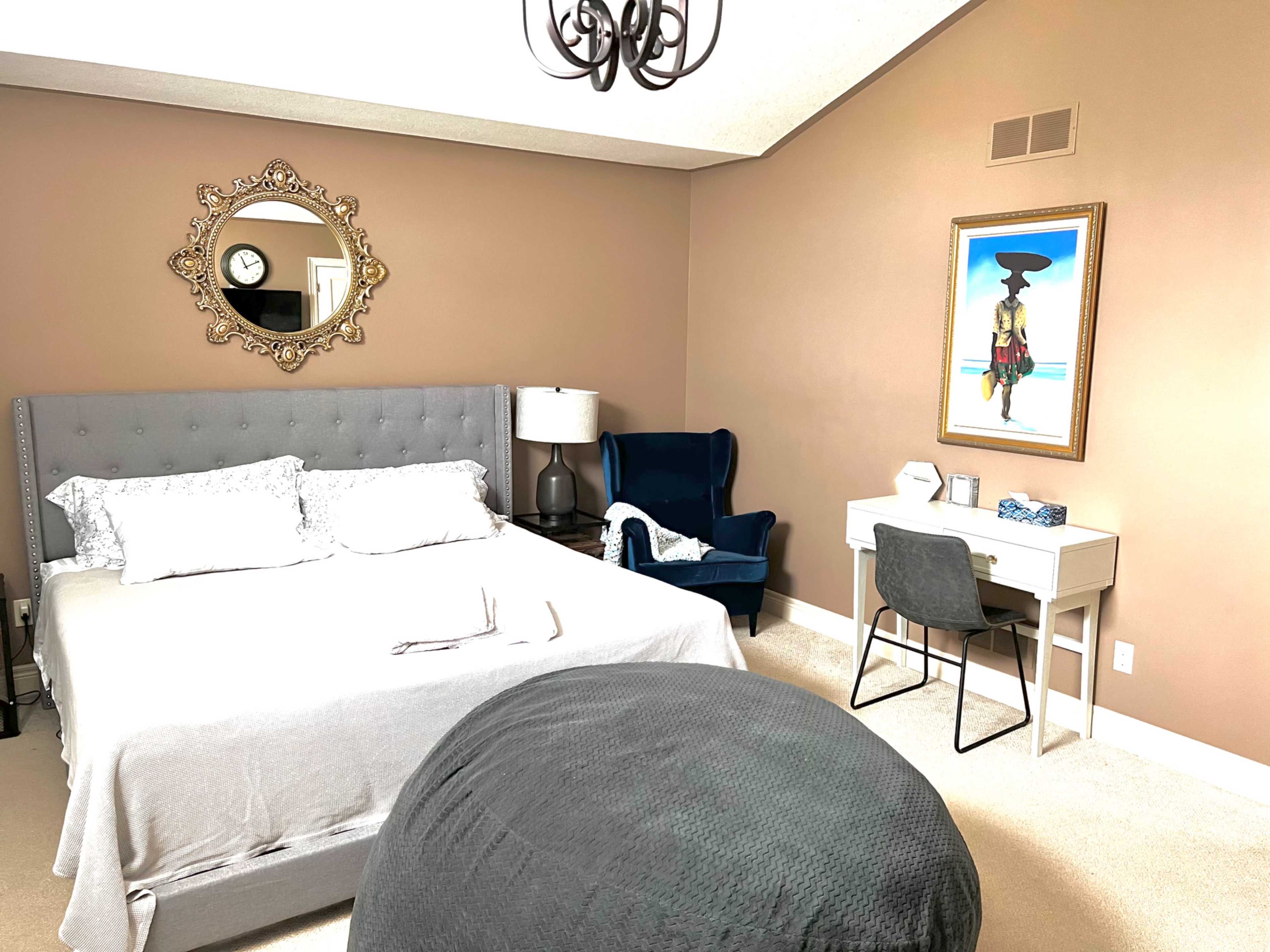 The image depicts a well-decorated bedroom featuring a gray bed, a decorative mirror, a blue armchair, and a small desk with a chair.