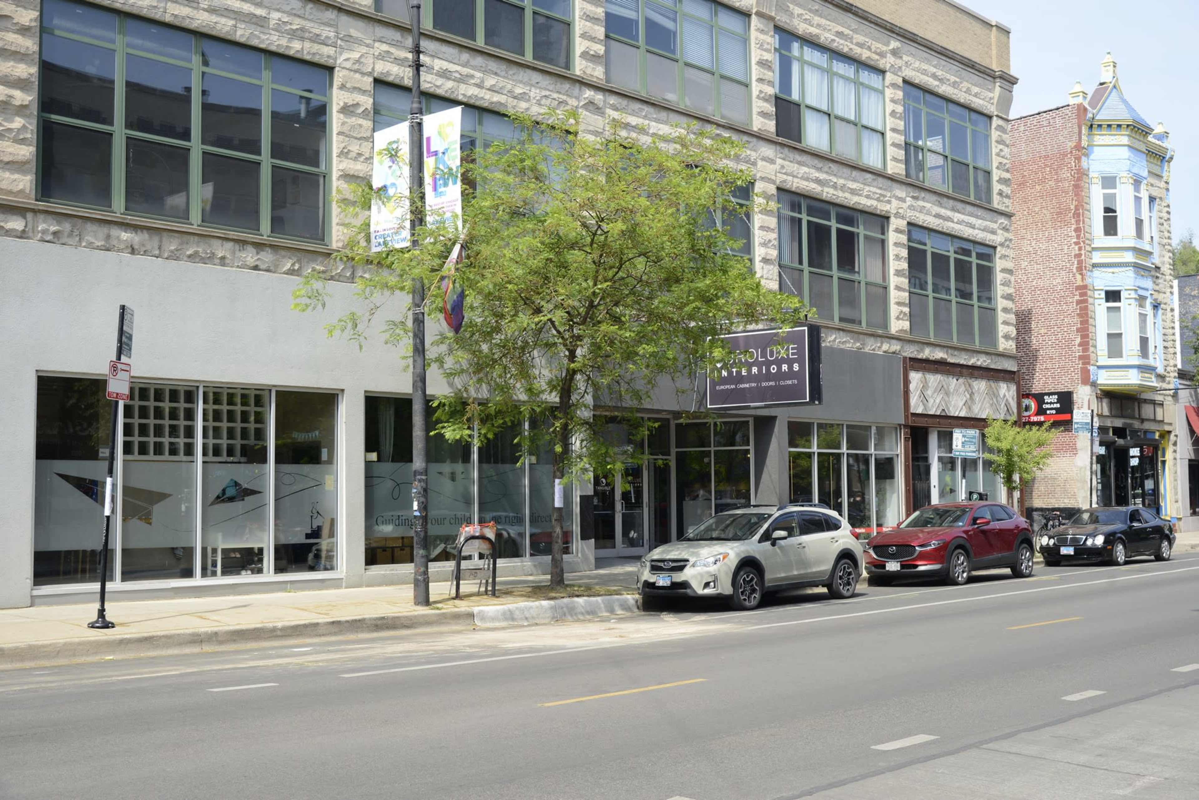 A street view of a multi-story building with large windows, a tree in front, and several parked cars along the curb.