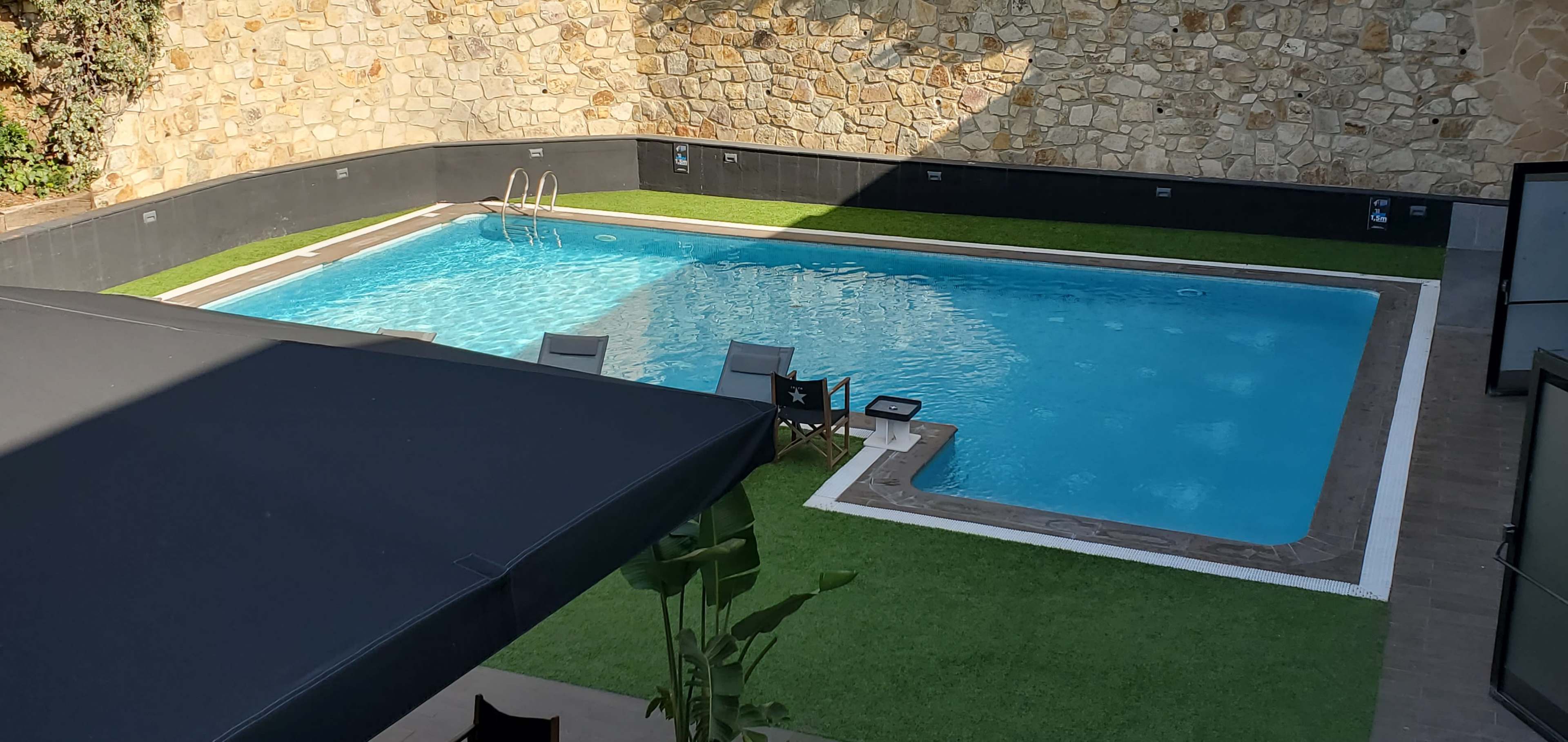 The image shows a rectangular swimming pool surrounded by artificial grass and a stone wall backdrop.