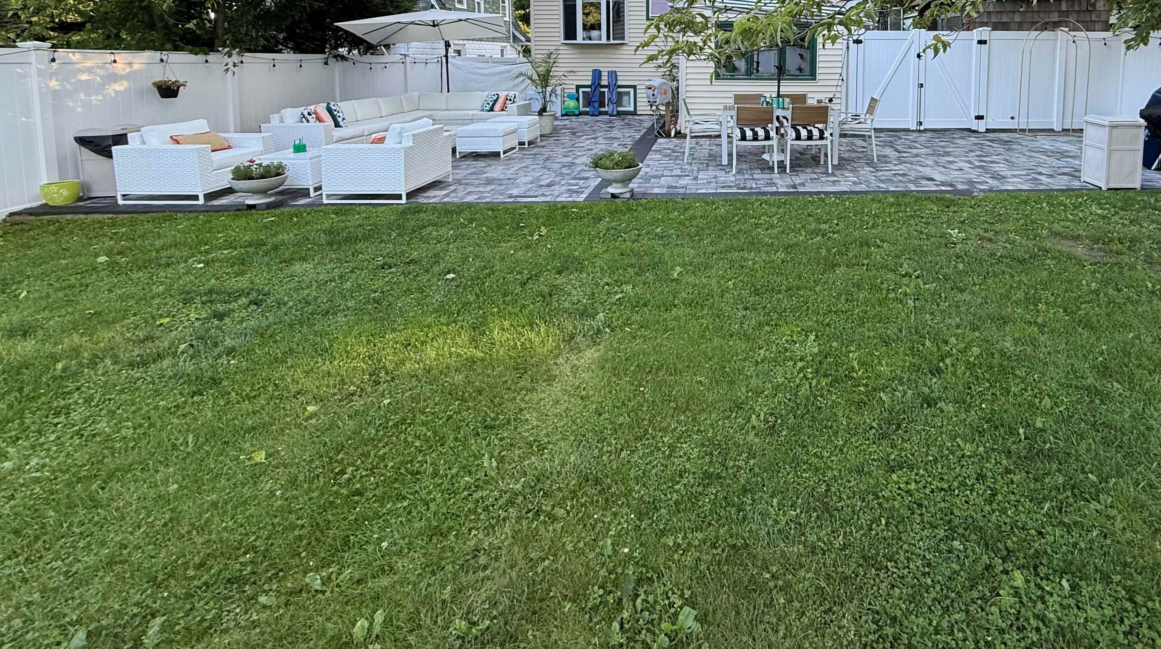 The image shows a backyard patio area with white outdoor furniture, a table, and a lawn in the foreground, surrounded by a white fence.