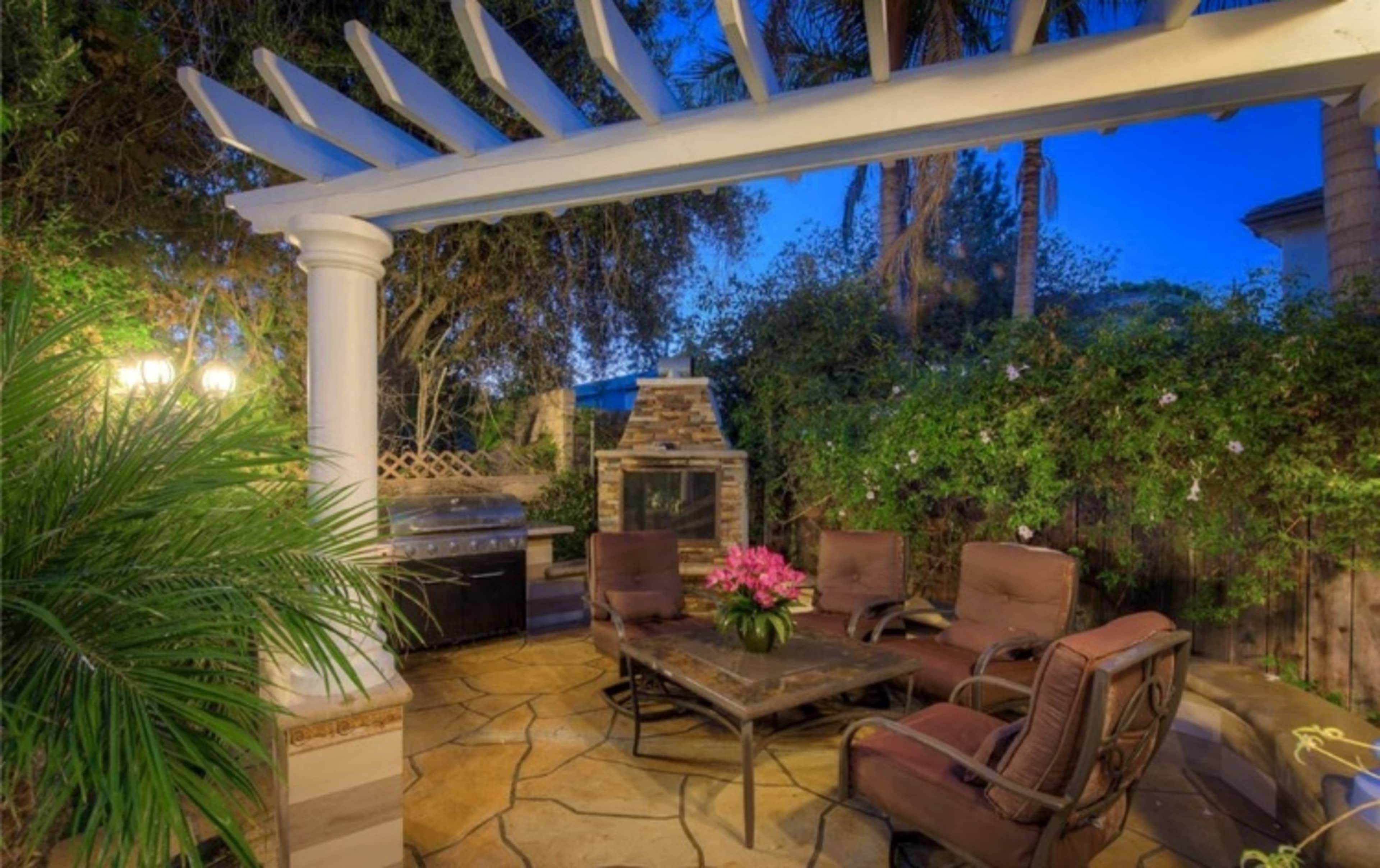 A shaded patio features a stone table surrounded by four chairs, with a barbecue grill and stone fireplace visible nearby.