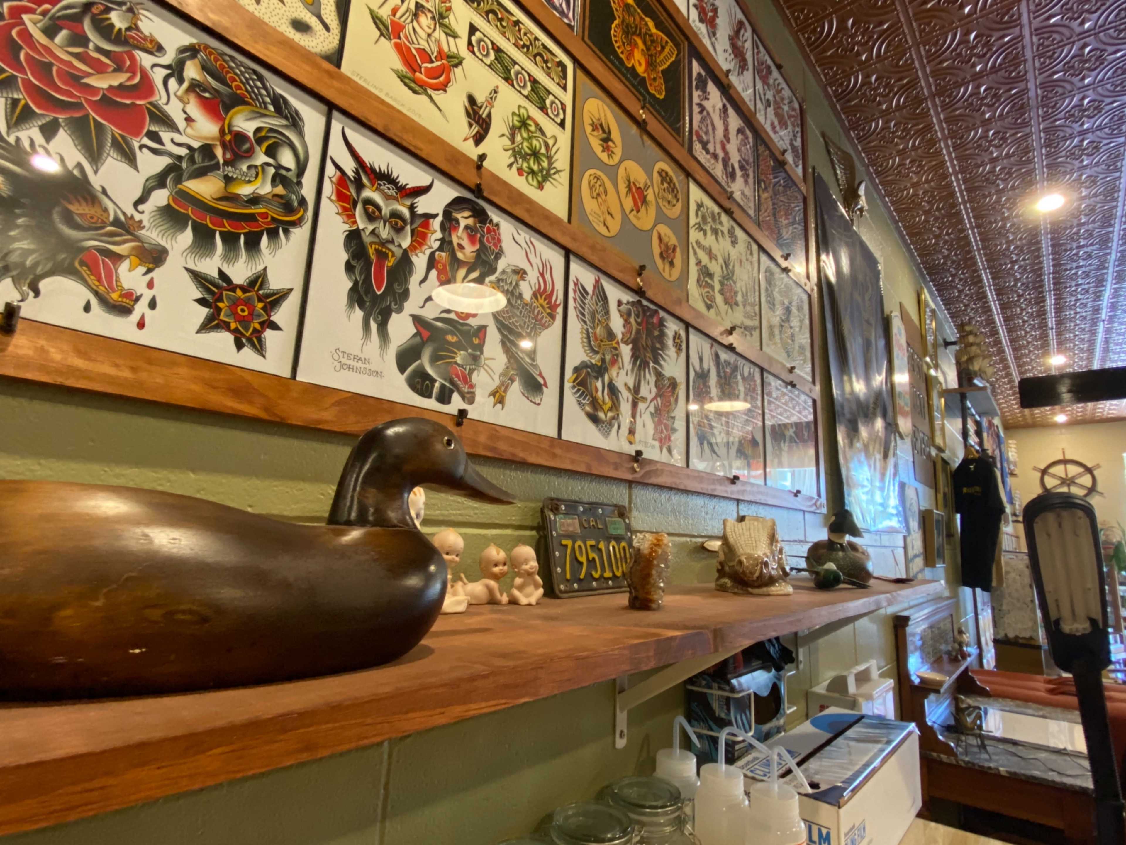 The image shows a wooden shelf displaying various decorative items, including a carved duck, small figurines, and glass containers, alongside a wall adorned with tattoo flash art.