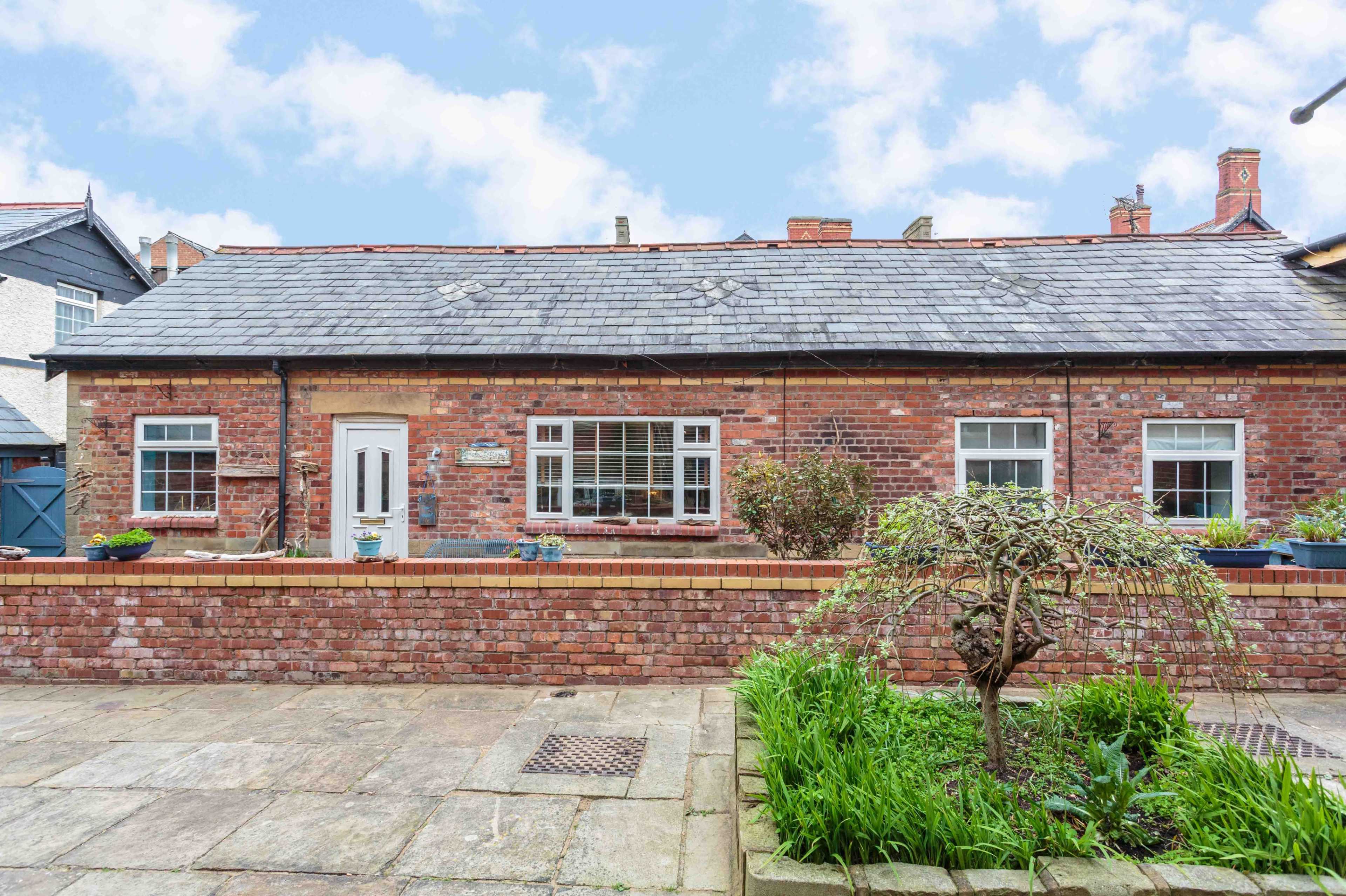 A single-story brick cottage with a slate roof and a landscaped front garden featuring plants and flowers.