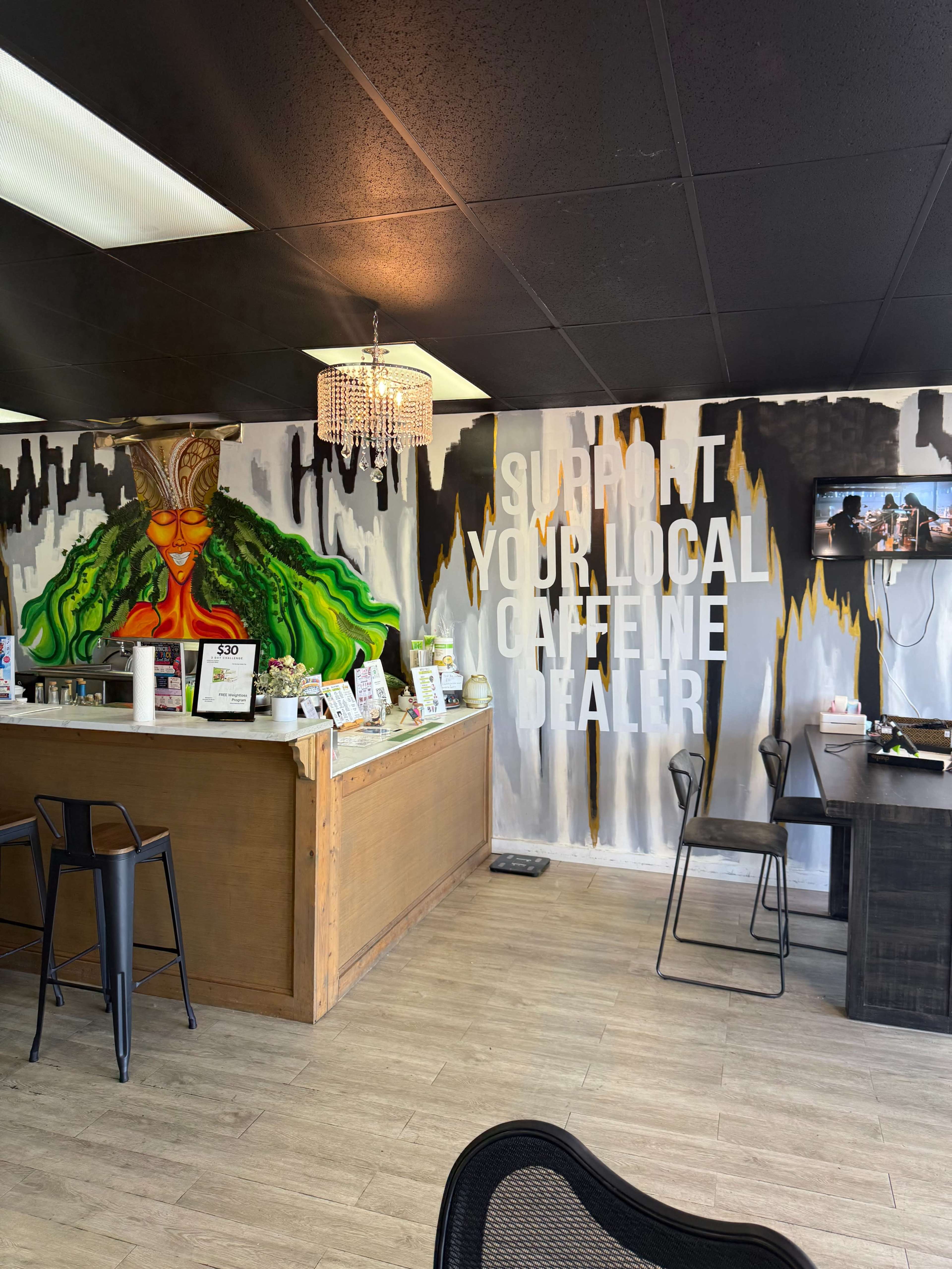 The interior of a coffee shop features a wooden counter, a vibrant mural of a woman with green hair, and a wall with the slogan "Support Your Local Caffeine Dealer."