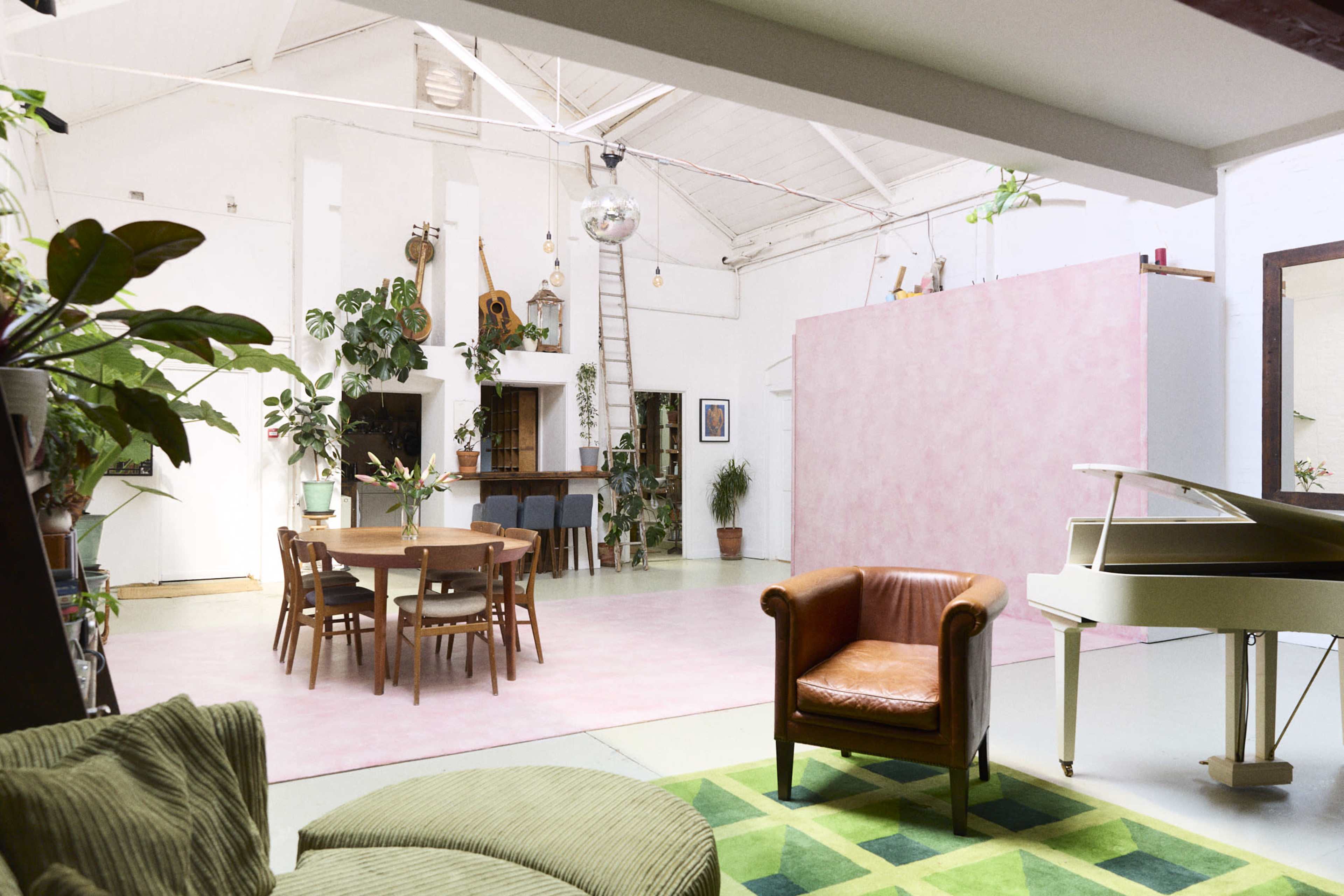 A spacious, open interior with a round dining table, a brown leather armchair, a white piano, and various plants against a backdrop of pink and white walls.