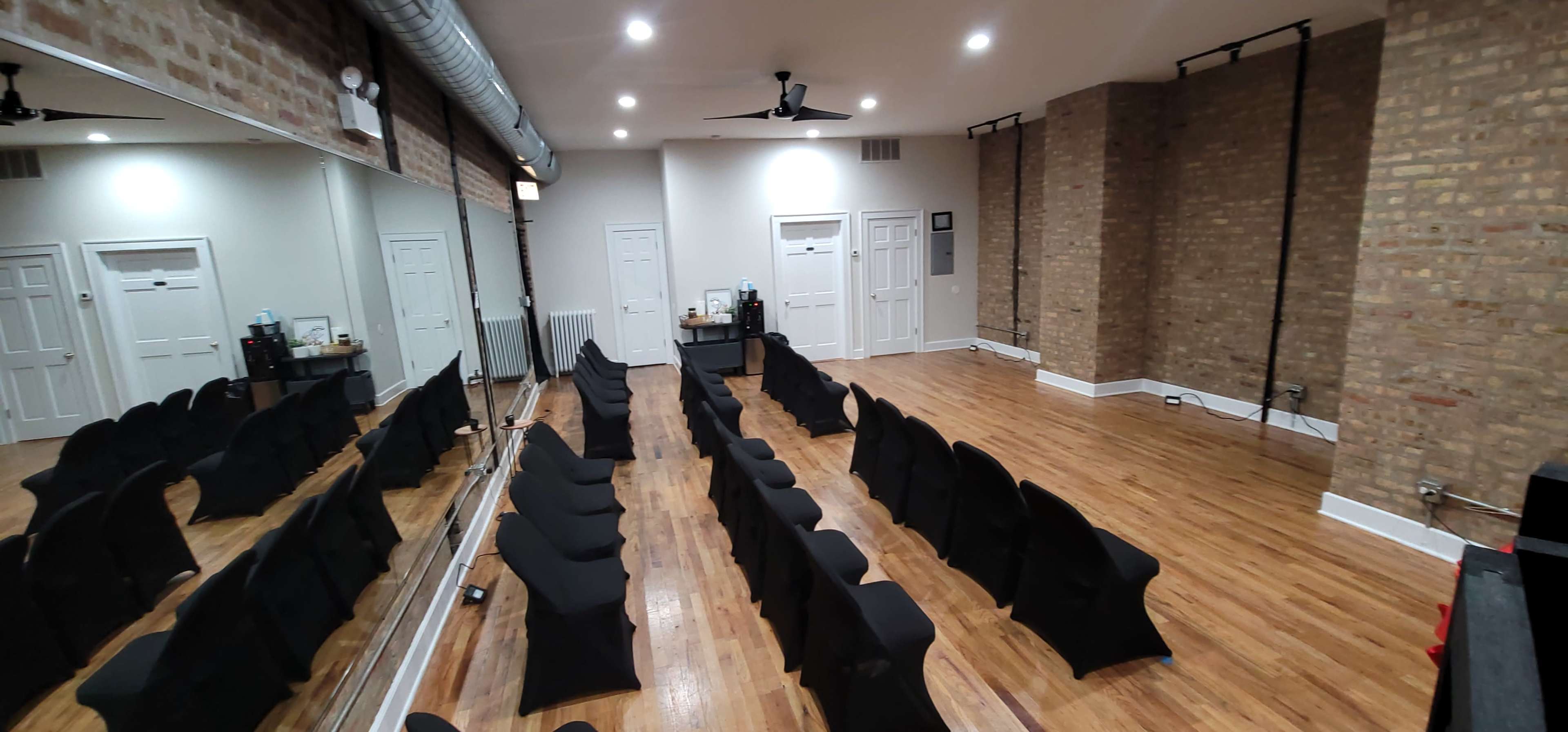 The image shows a spacious room with wooden flooring, featuring rows of black-covered chairs and a large mirror on one wall.