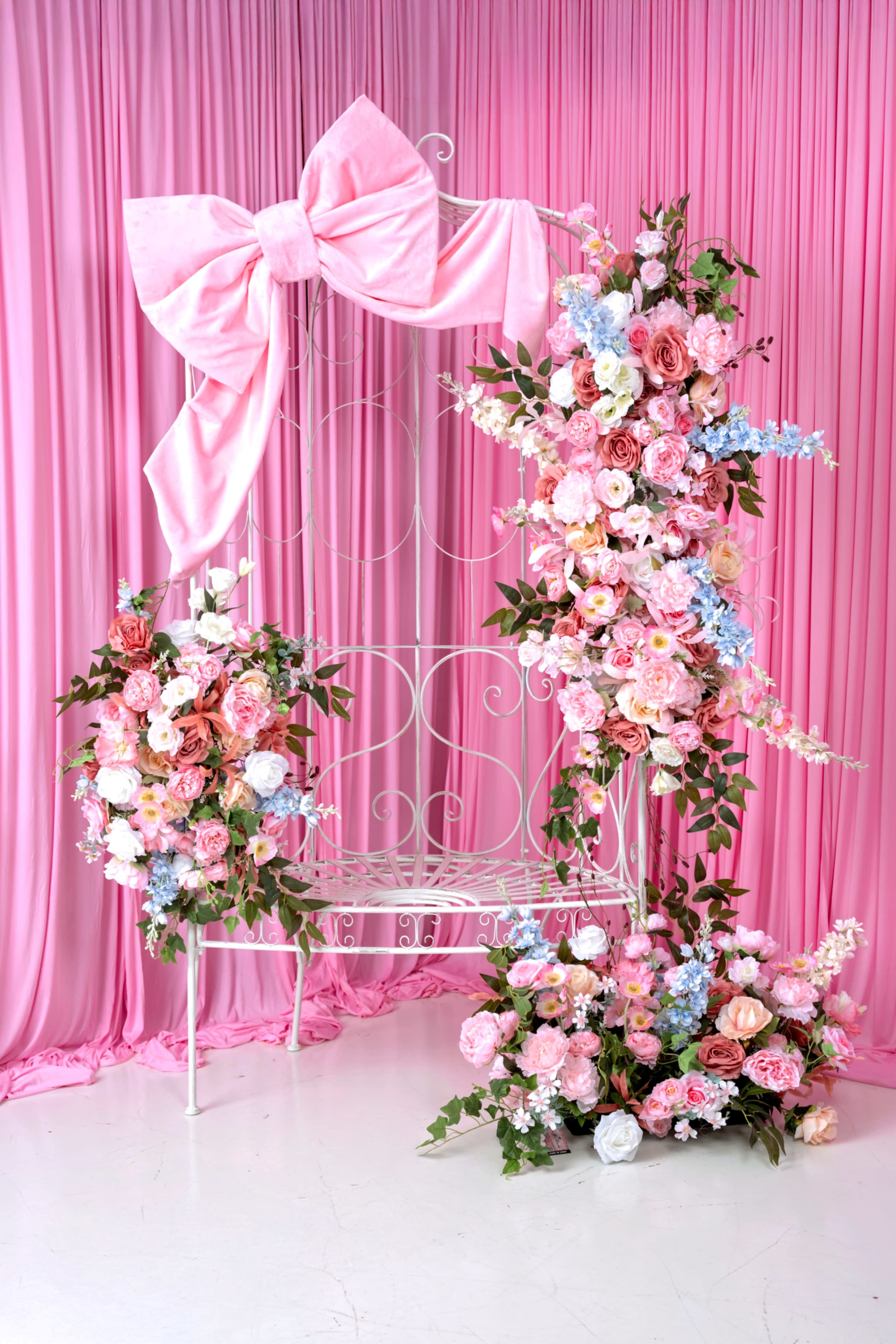 The scene features a decorative backdrop with a large pink bow, surrounded by arrangements of colorful artificial flowers, set against a pink curtain.
