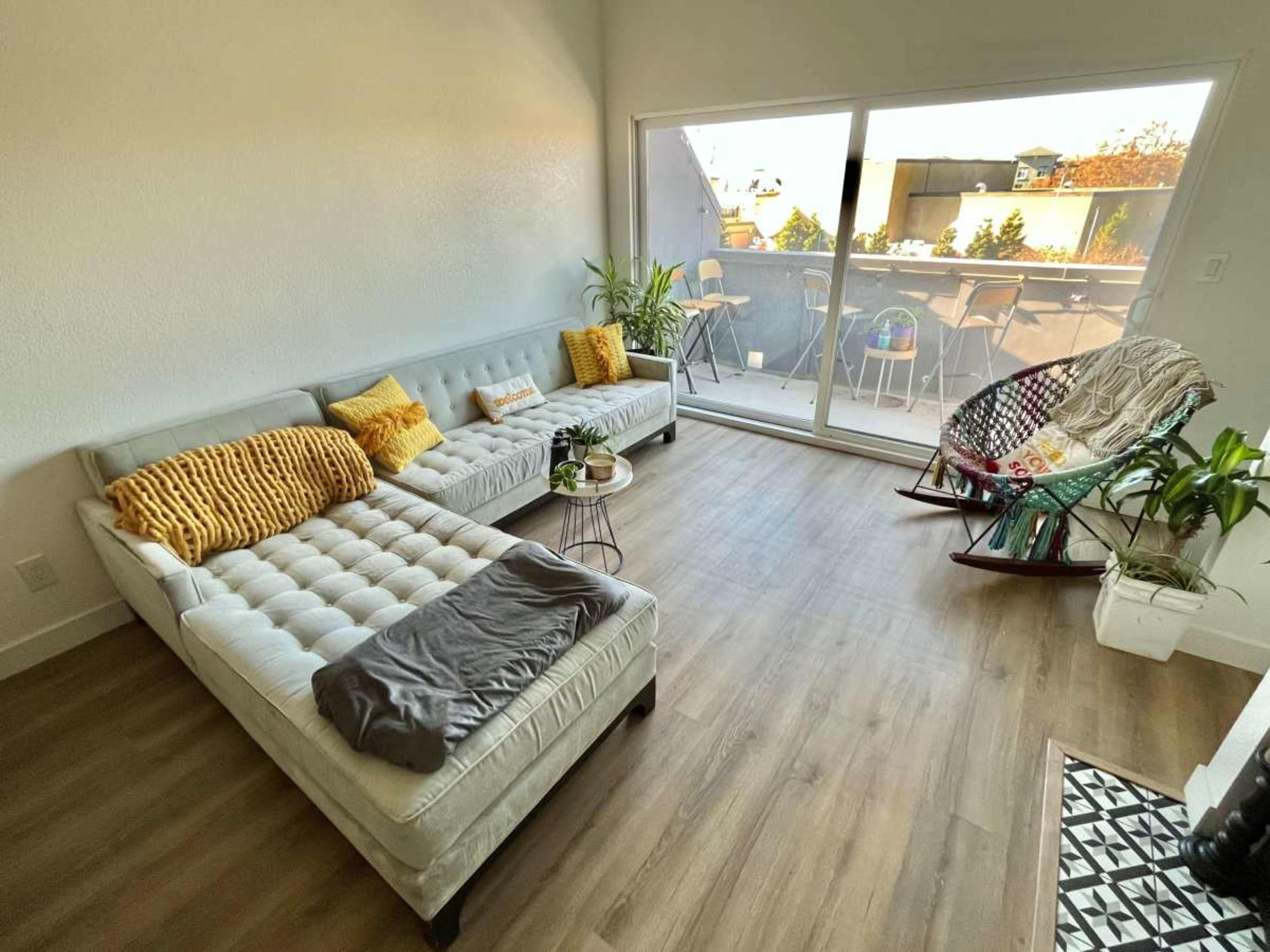 A light-filled living room features a gray sectional sofa, a rocking chair, potted plants, and large sliding glass doors leading to a balcony.