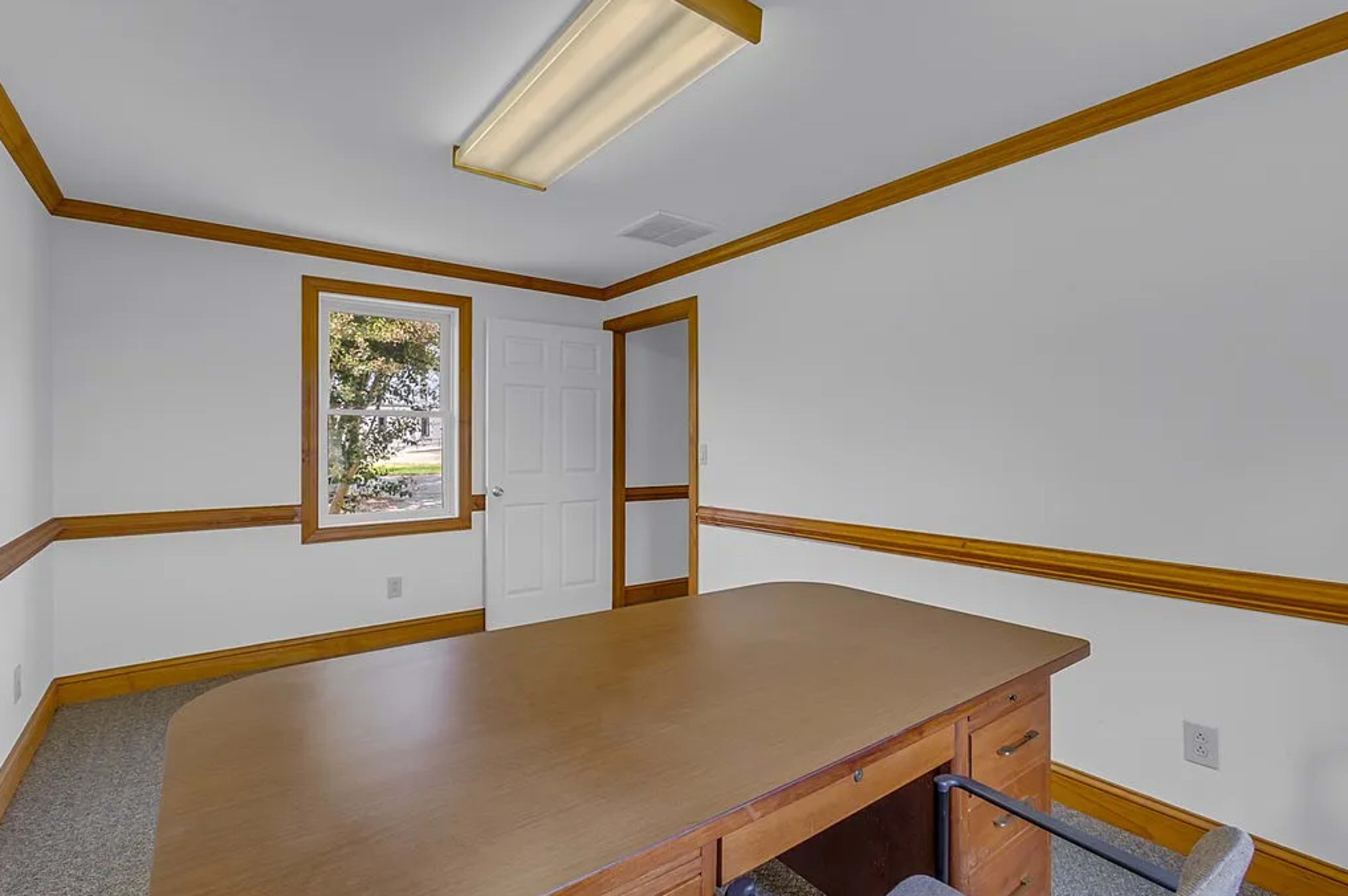 The image shows a small office room featuring a wooden table, a single chair, and a window overlooking an outdoor area.