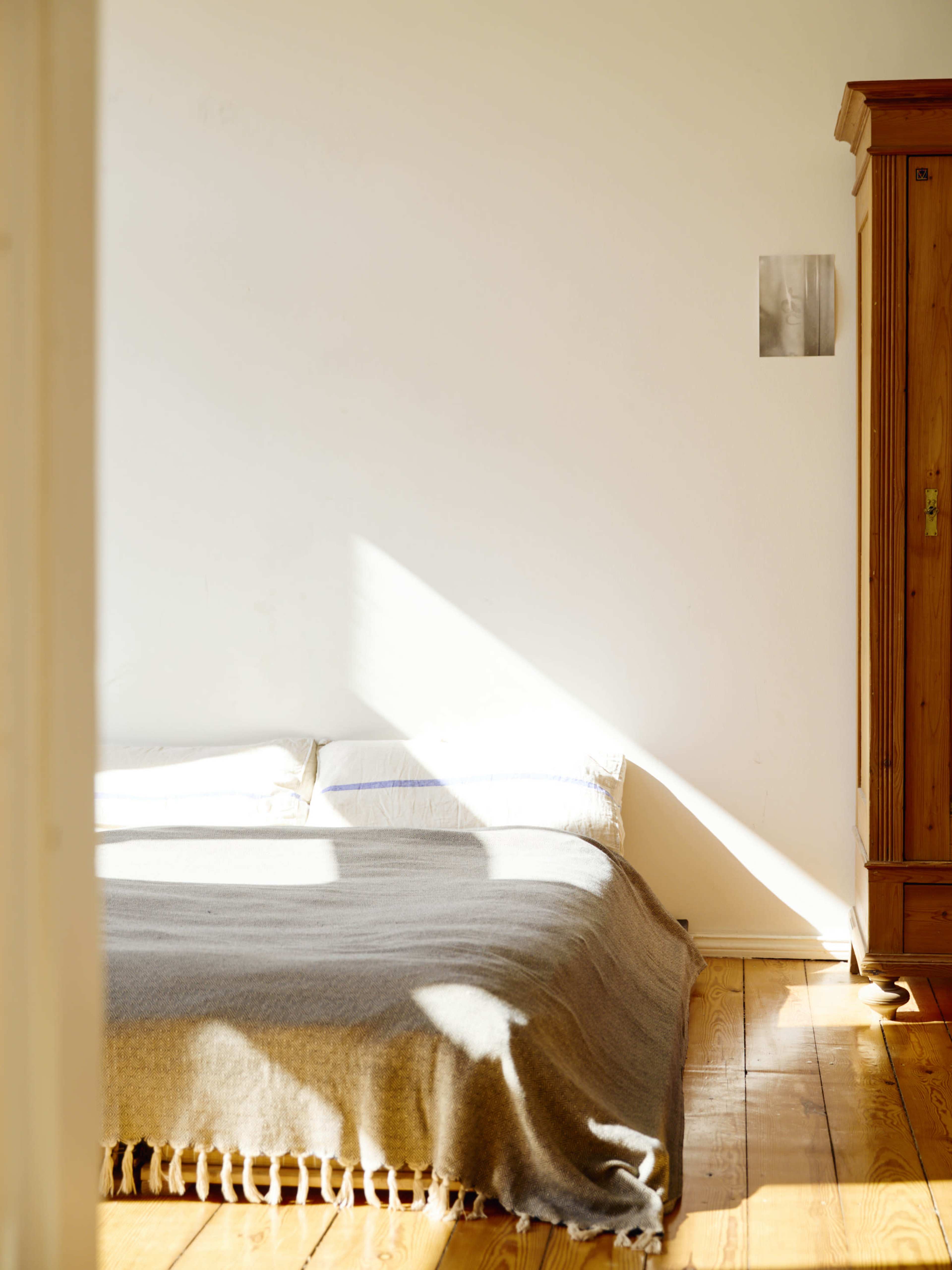 A bed with a gray blanket is positioned against a wall, illuminated by sunlight, with a wooden cabinet nearby.