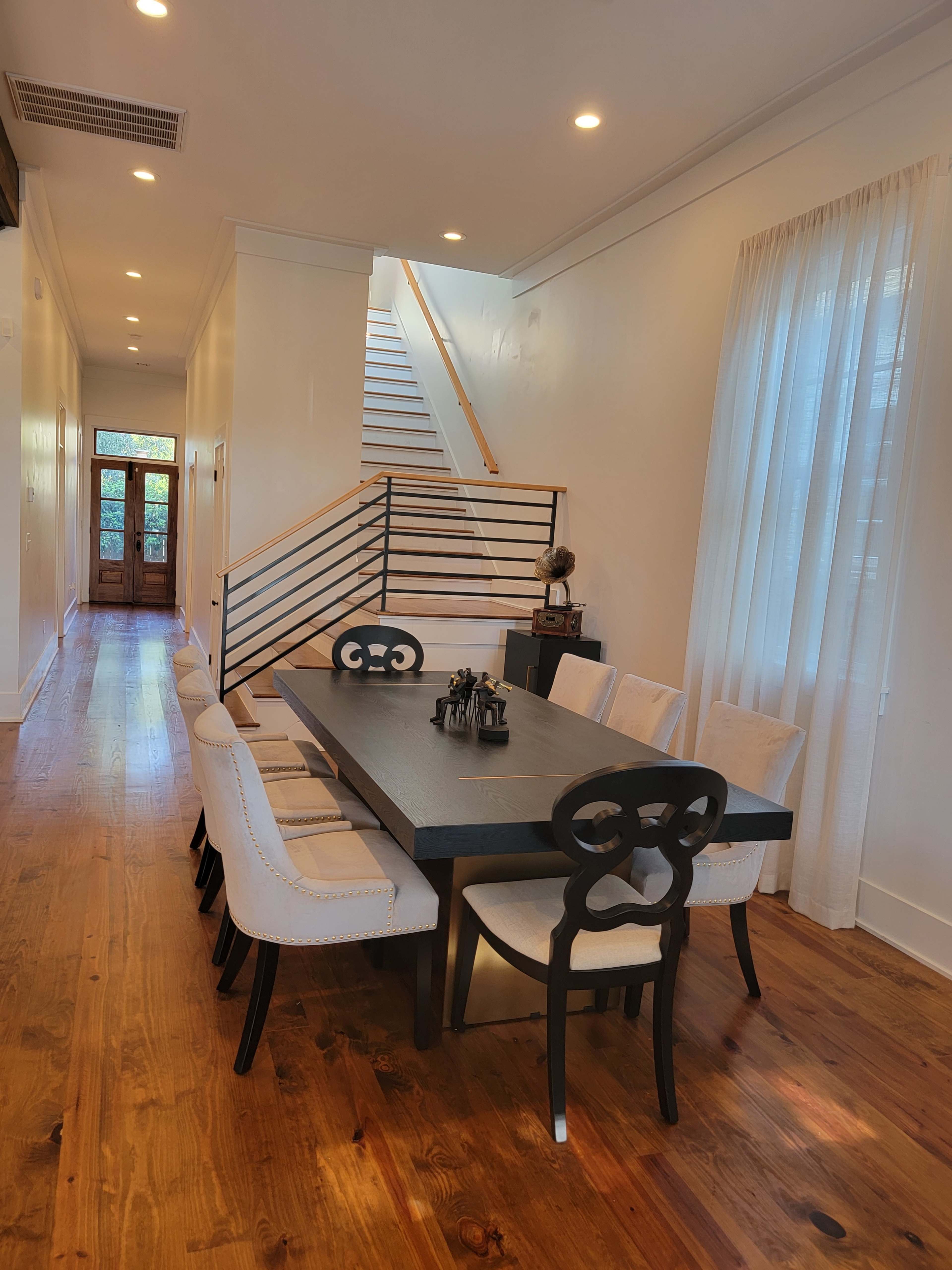 A well-lit dining area with a long black table surrounded by upholstered chairs, leading towards a staircase and an entryway with wooden doors.