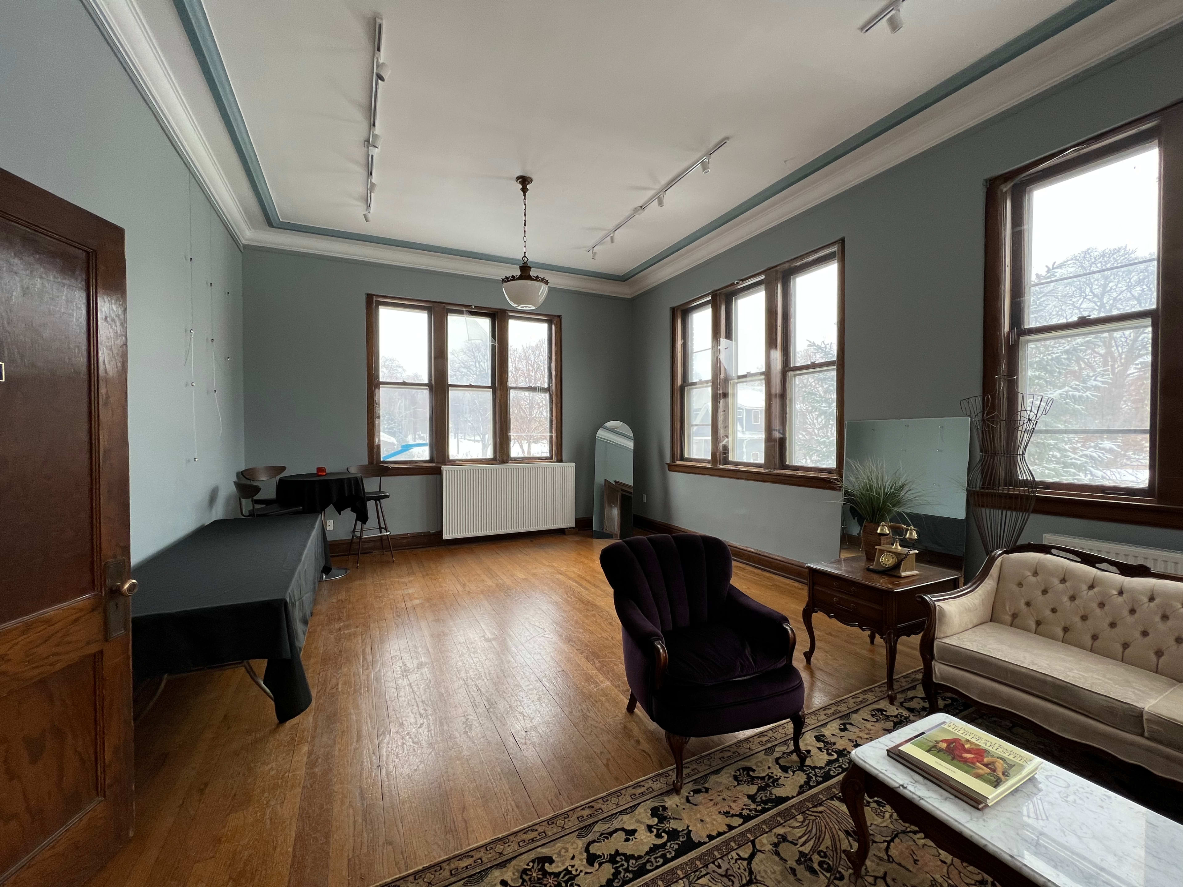 A spacious, empty room with large windows, hardwood floors, and a mixture of furniture including a velvet armchair and a couch.