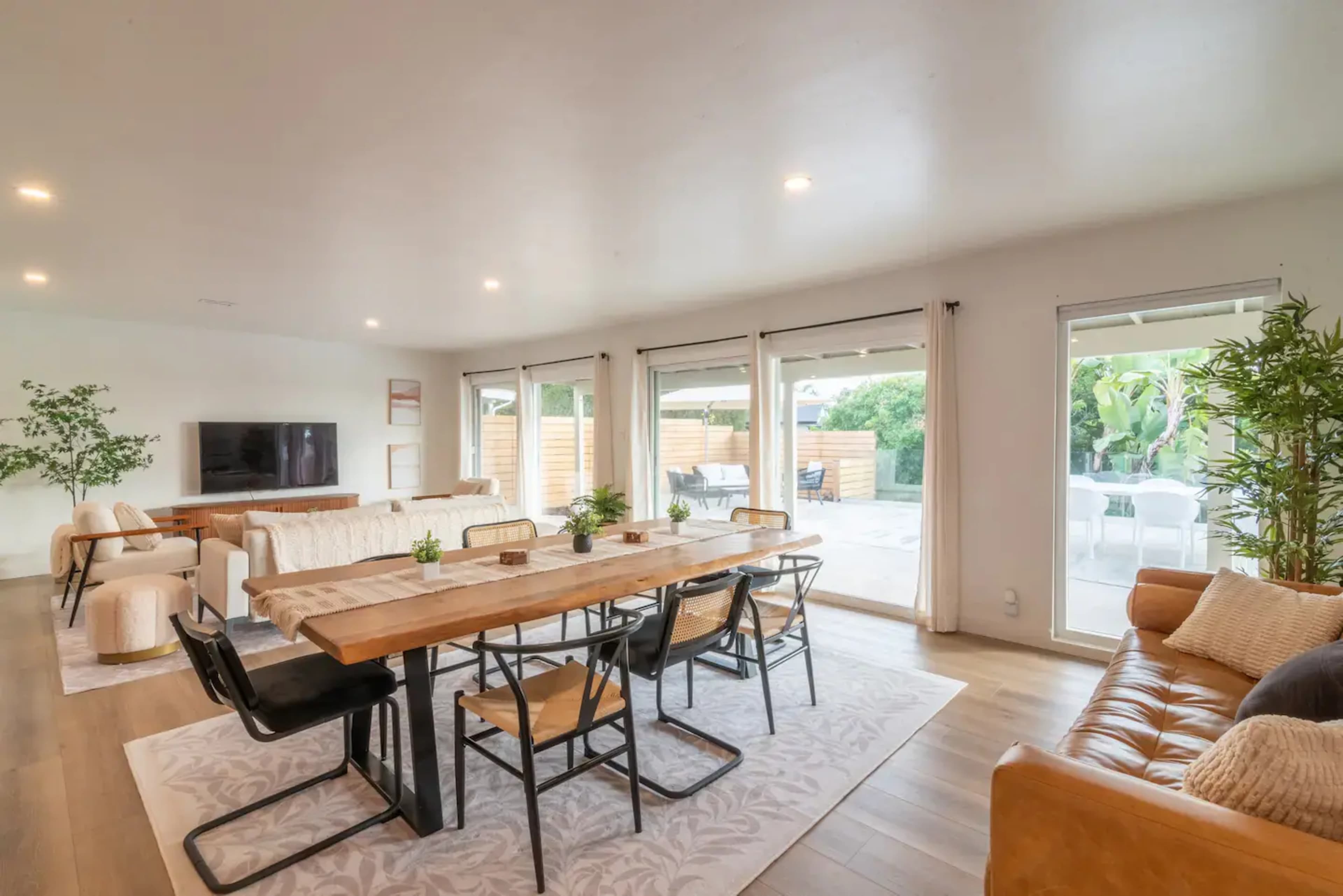 A spacious living and dining area features a wooden table surrounded by black chairs, an exposed brick wall, and large windows leading to an outdoor space.