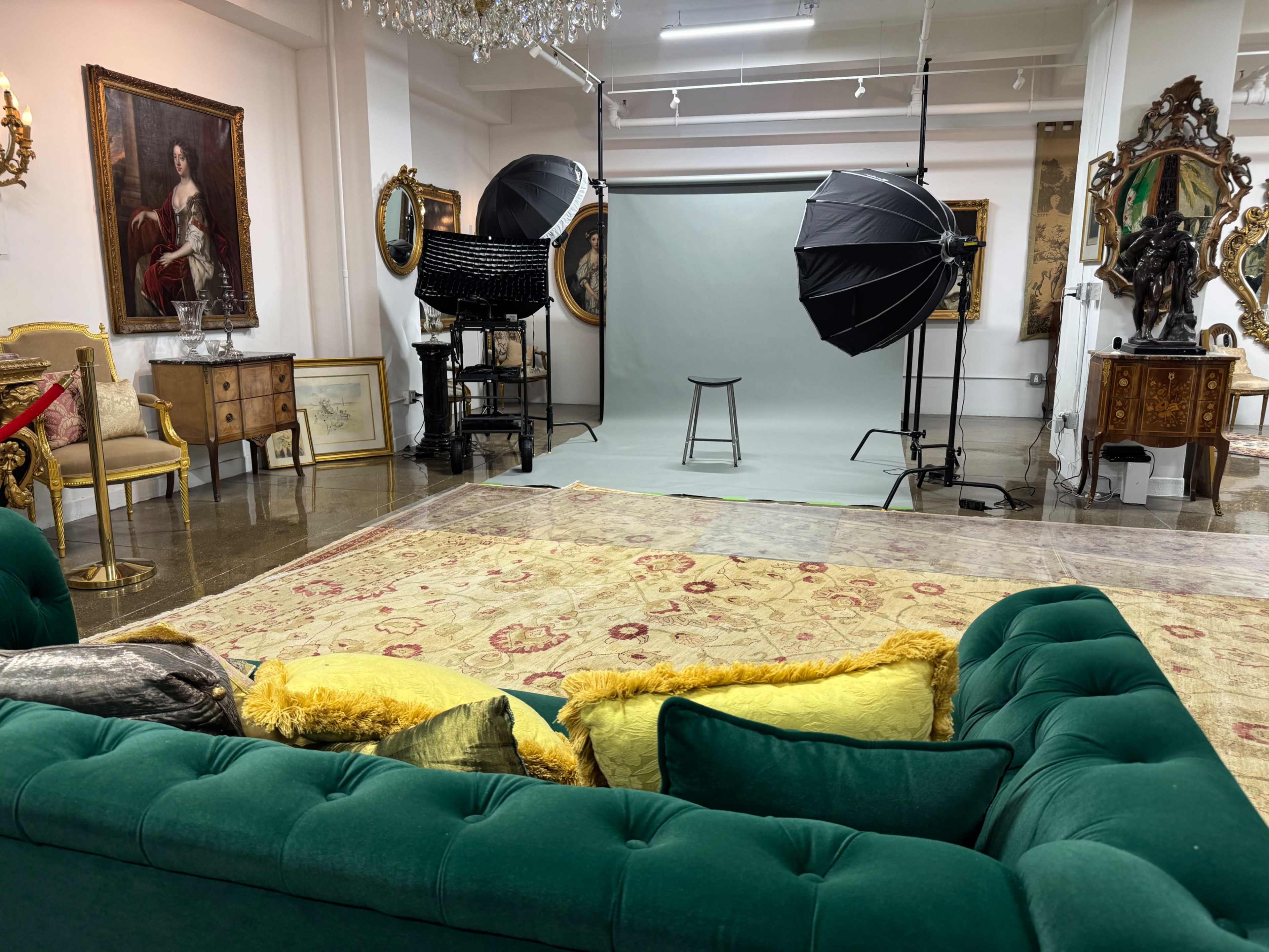 The image shows a chic interior space featuring a green sofa, a light-colored backdrop with studio lighting setup, and various ornate furnishings and artwork on the walls.