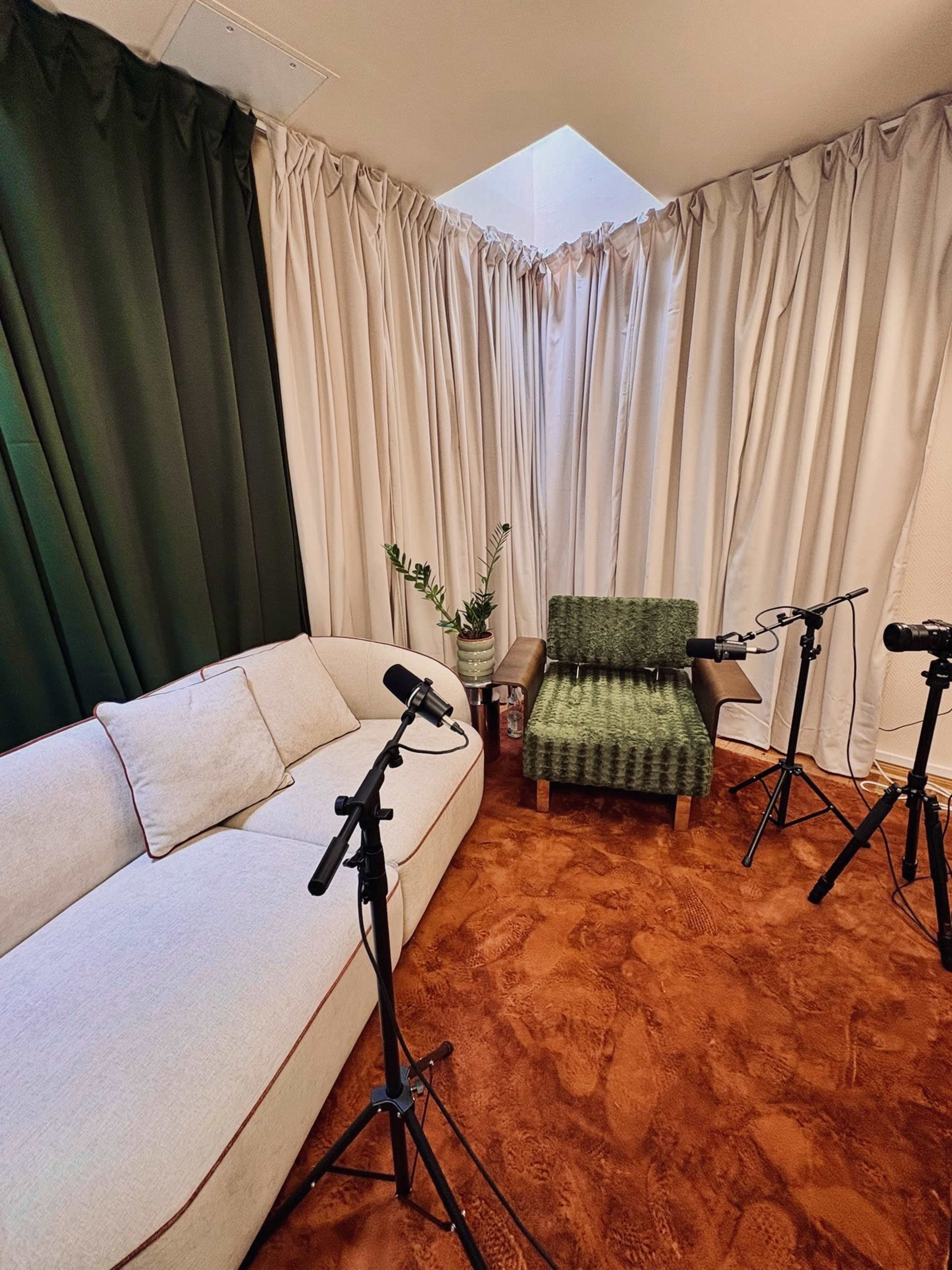 The image shows a small room with a white sofa, a green upholstered chair, and two microphones on stands positioned around a potted plant.