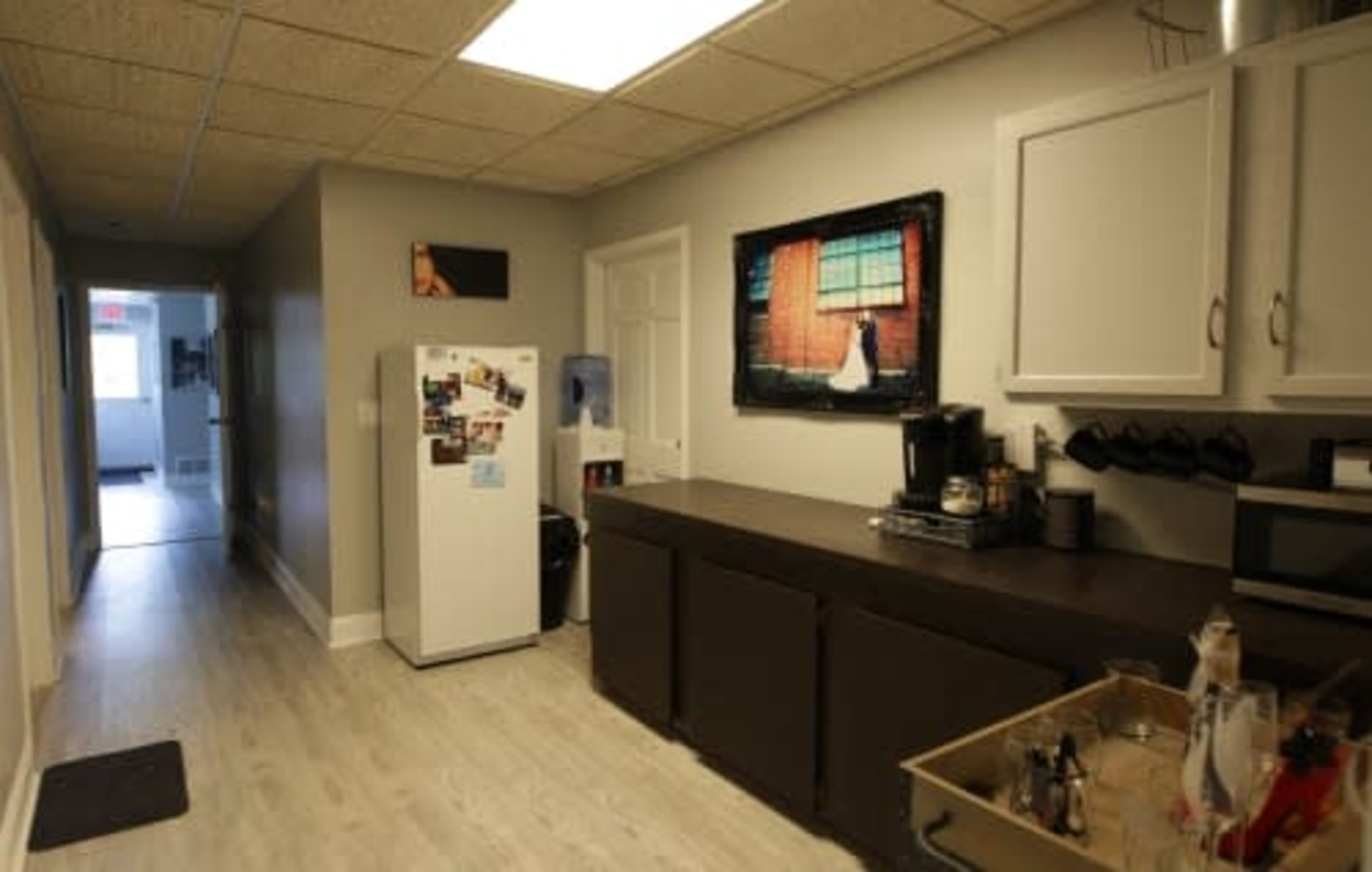 A kitchen with a refrigerator, countertop, coffee maker, and a wall-mounted television displaying a scene, along with a hallway leading to another room.