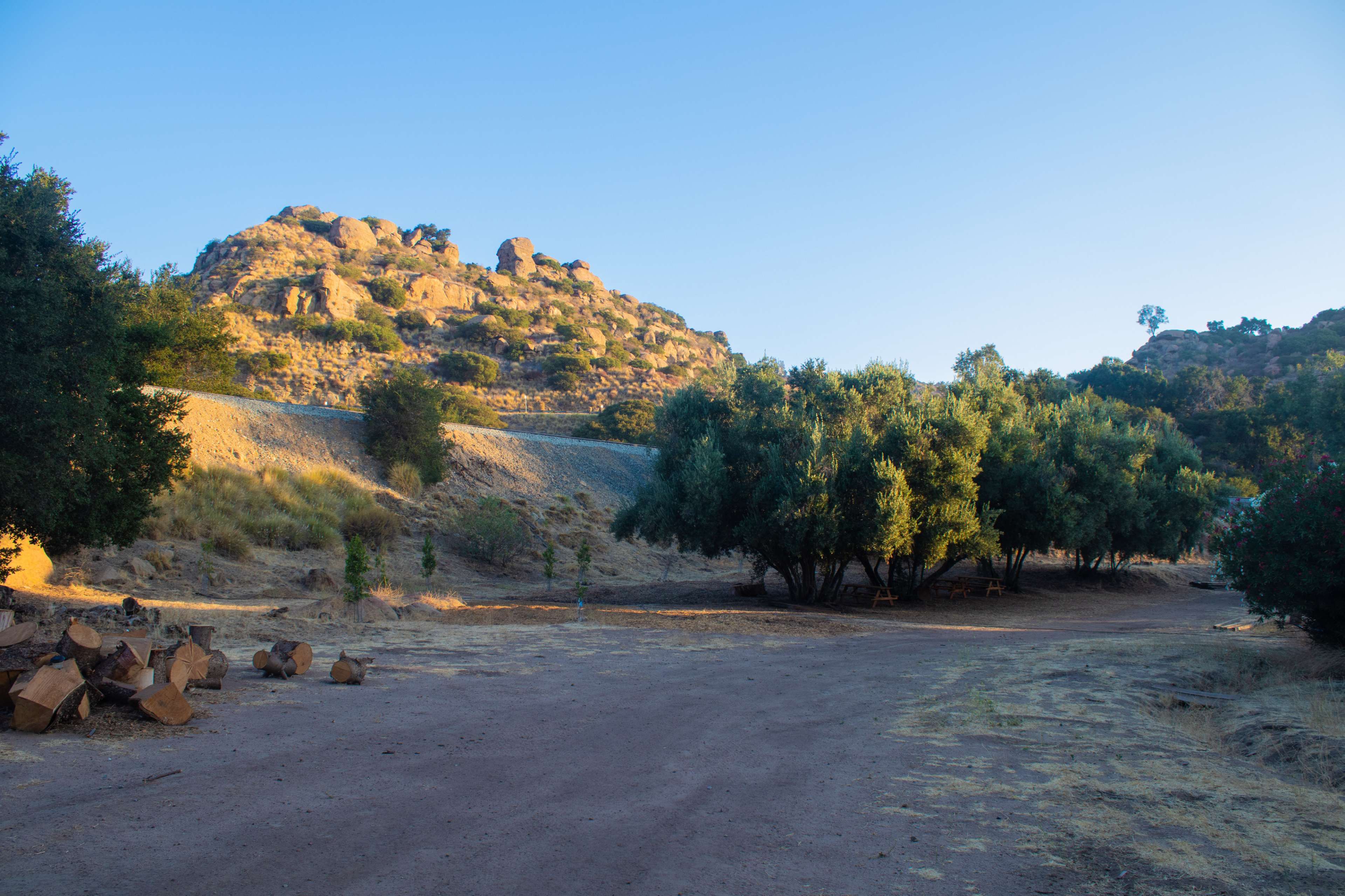 A dirt path leads through a sparse landscape with a rocky hill and clusters of trees under a clear sky.
