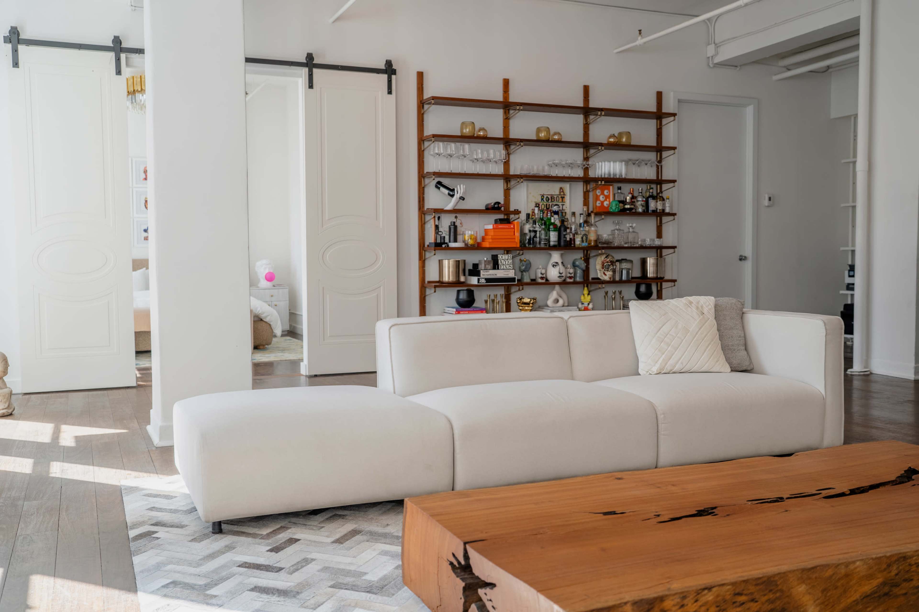 The image shows a modern living room with a white sectional sofa, a wooden coffee table, and a shelving unit displaying various decorative items and glassware.
