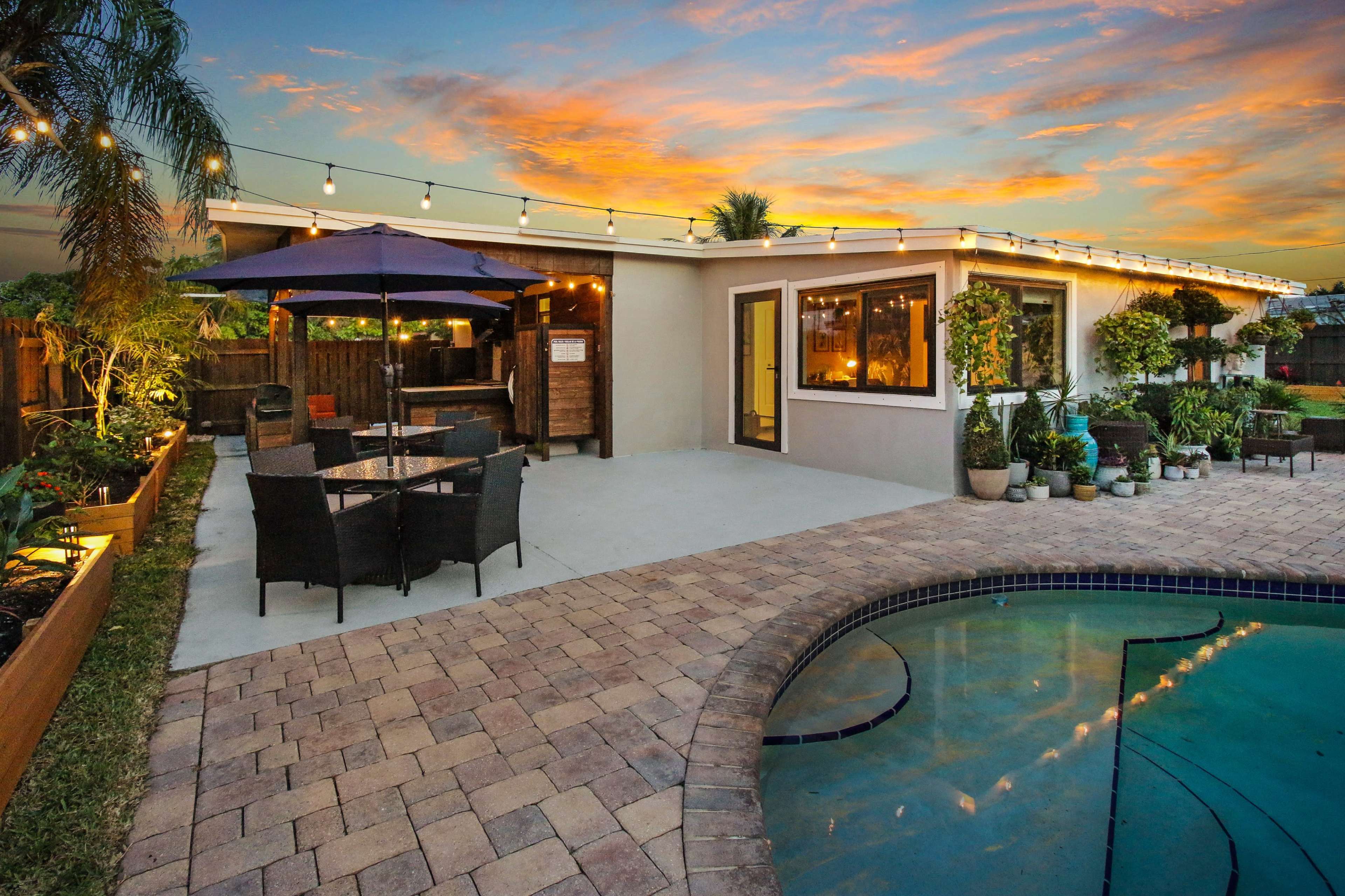 A backyard features a swimming pool, a patio with outdoor seating, and string lights overhead against a sunset sky.