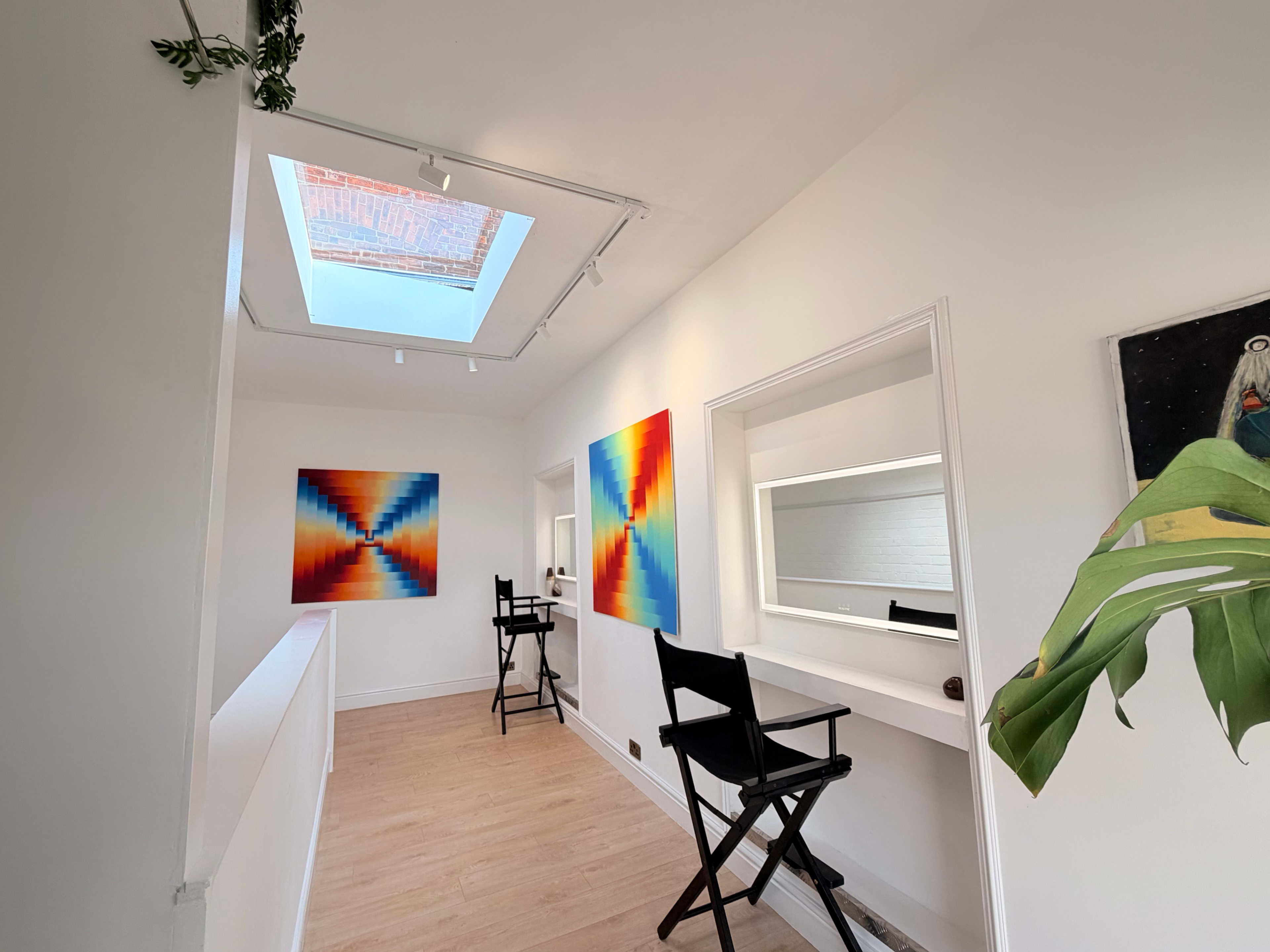 A brightly lit hallway features two vibrant abstract paintings on the walls, a large skylight above, and two director's chairs positioned near a mirror.