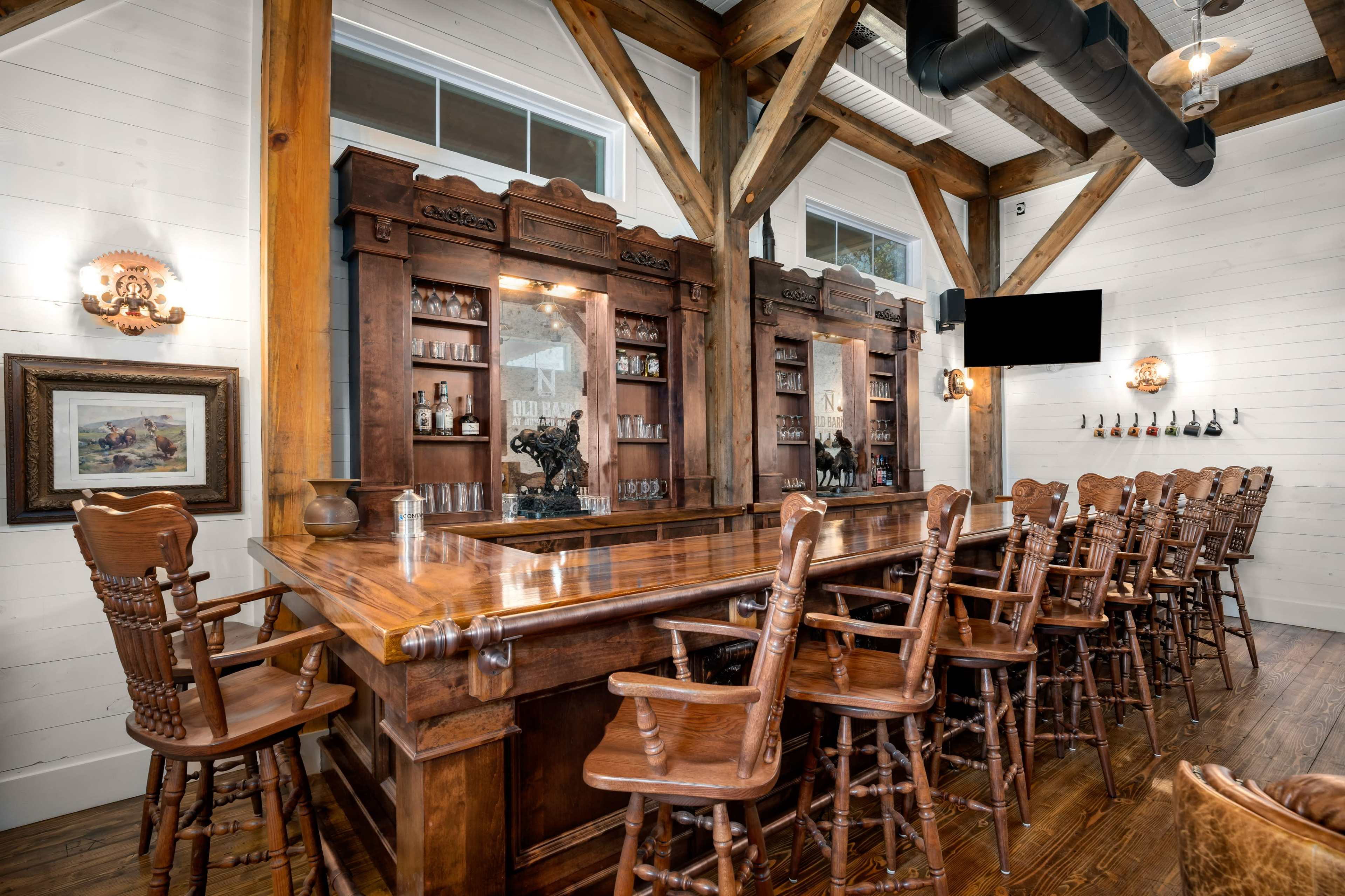 A wooden bar with high stools arranged around it, featuring a dark wooden backlit shelf displaying bottles and glasses, set in a room with exposed beams and large windows.