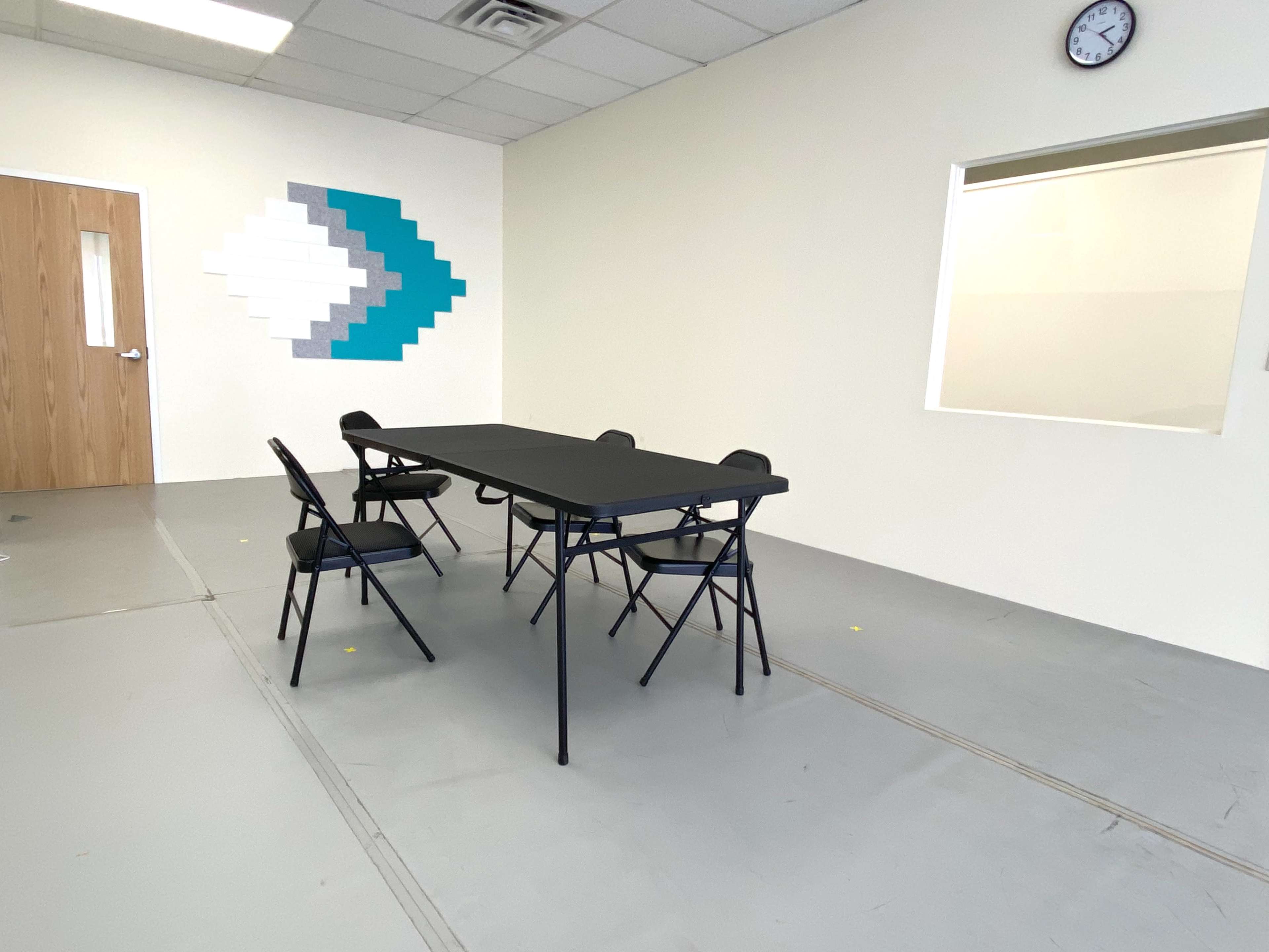 A rectangular table with four folding chairs is positioned in a room that features a wall decoration of teal and white geometric shapes.