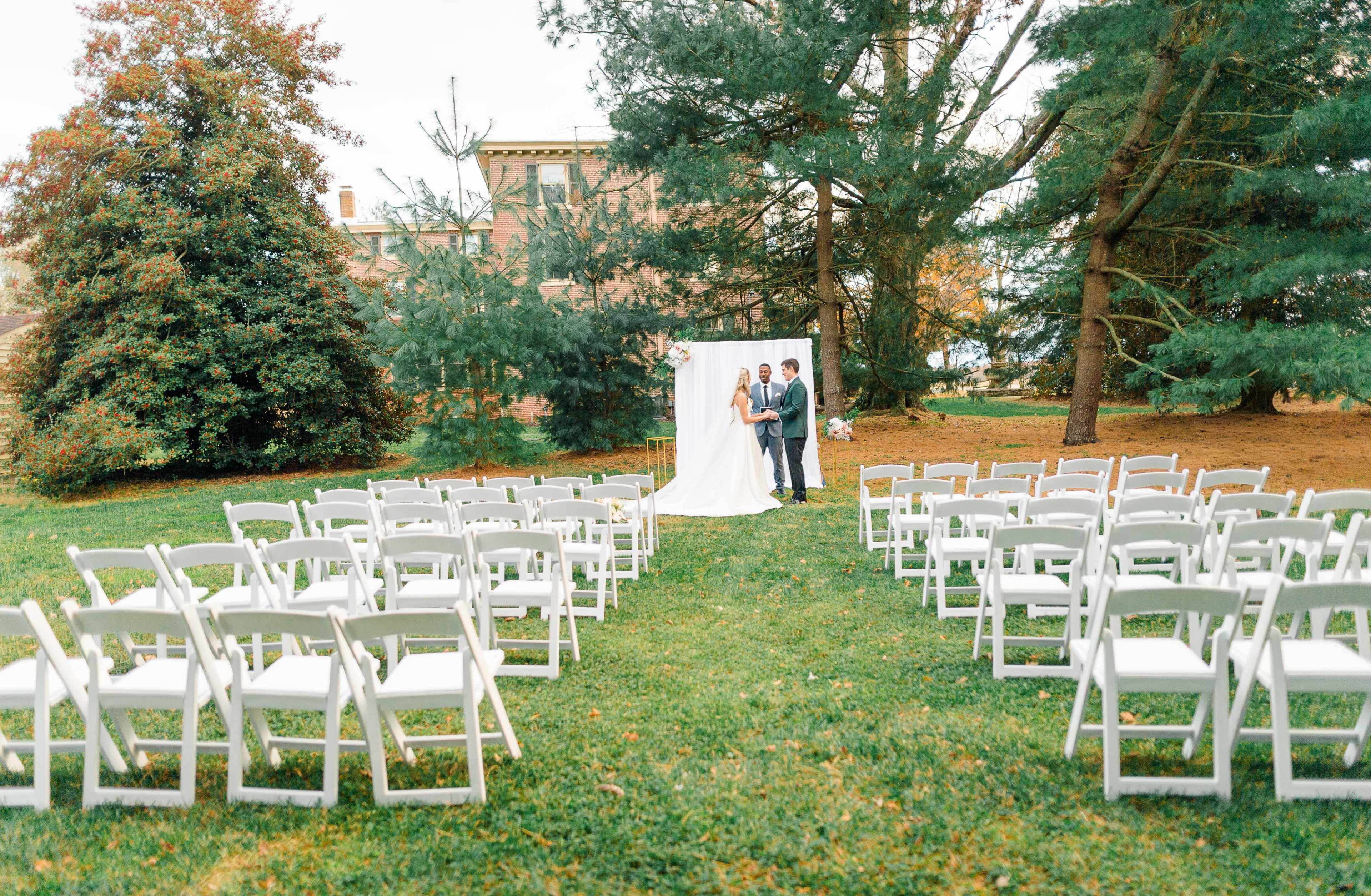 A wedding ceremony setup features white folding chairs arranged in rows facing a backdrop of white drapery among green trees.