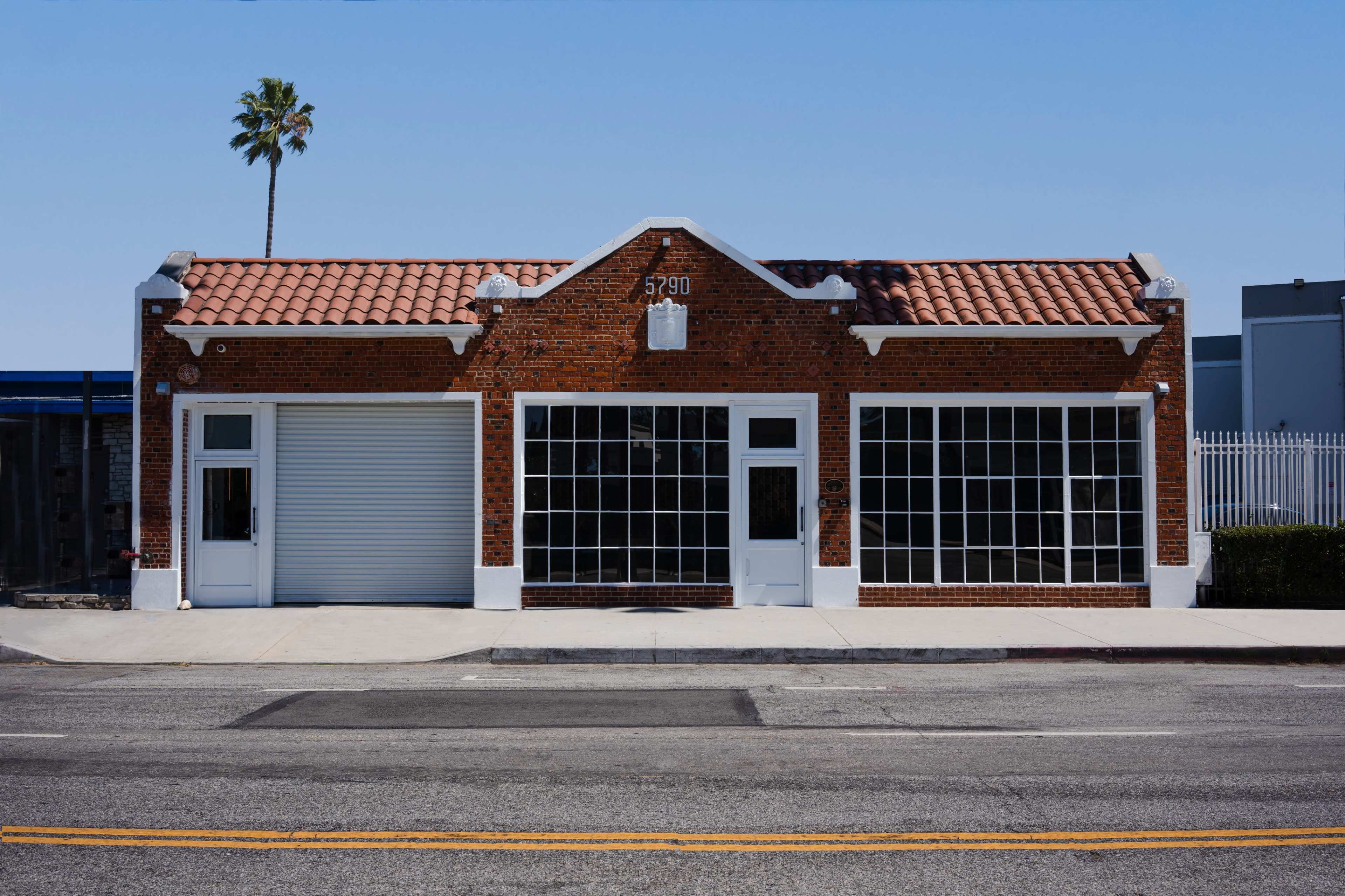 A brick building with a tiled roof features large windows and a roll-up garage door, located by the street under a clear blue sky.