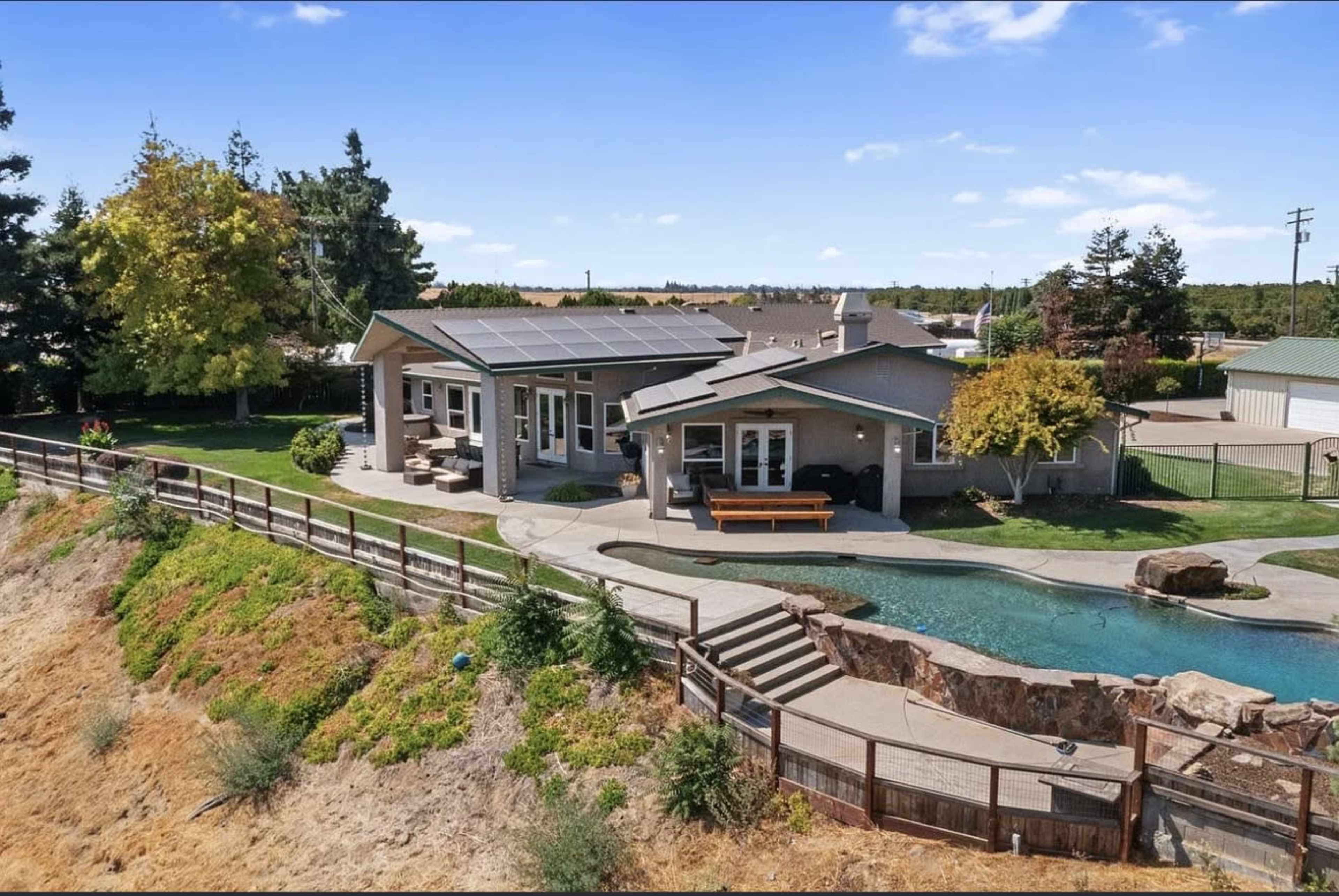 The image shows a modern single-story home with a swimming pool and landscaped yard, situated on a slope with a fence surrounding the property.