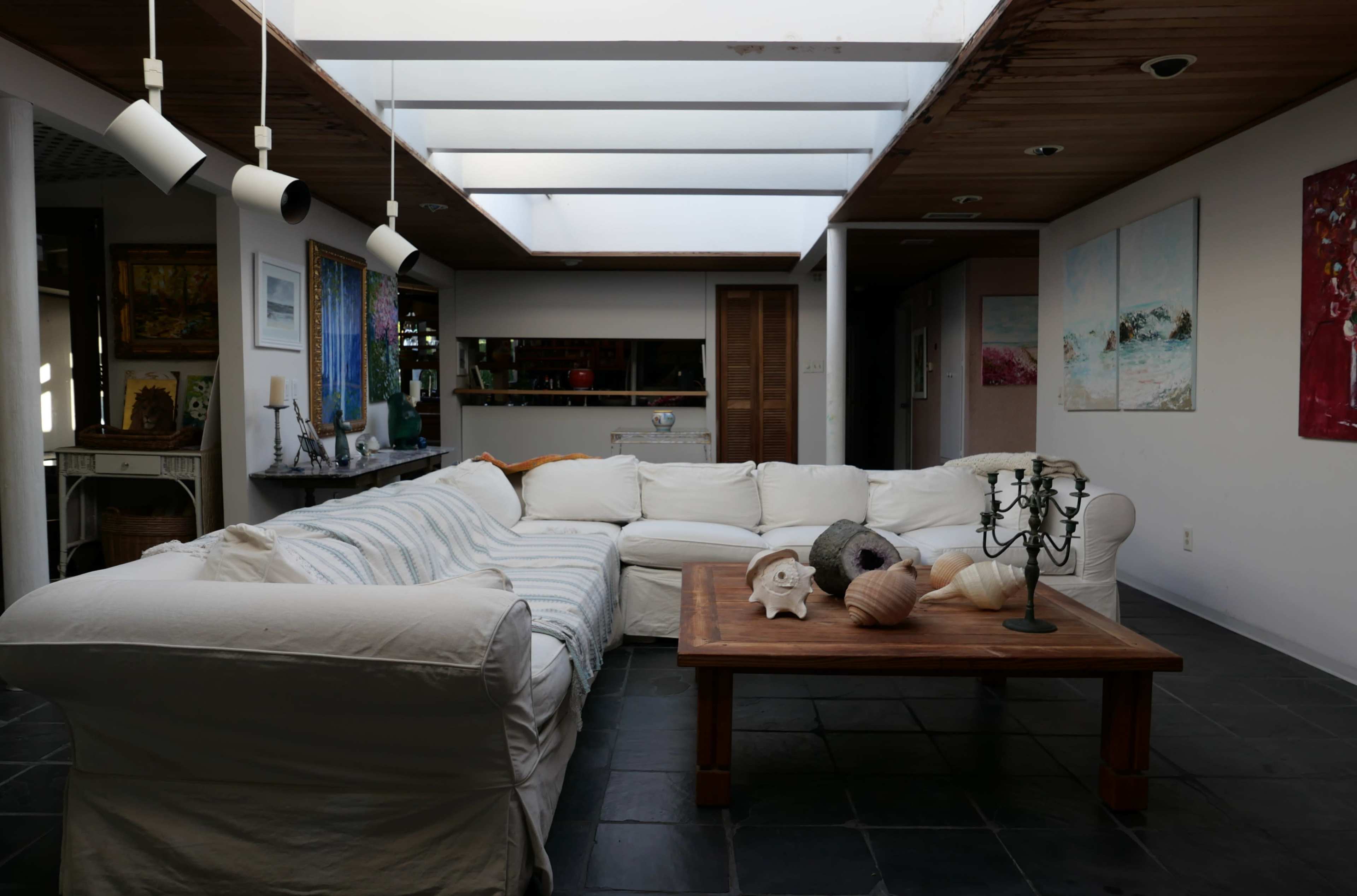 A modern living room with a white sectional sofa, a wooden coffee table adorned with decorative shells, and large windows allowing natural light to enter.