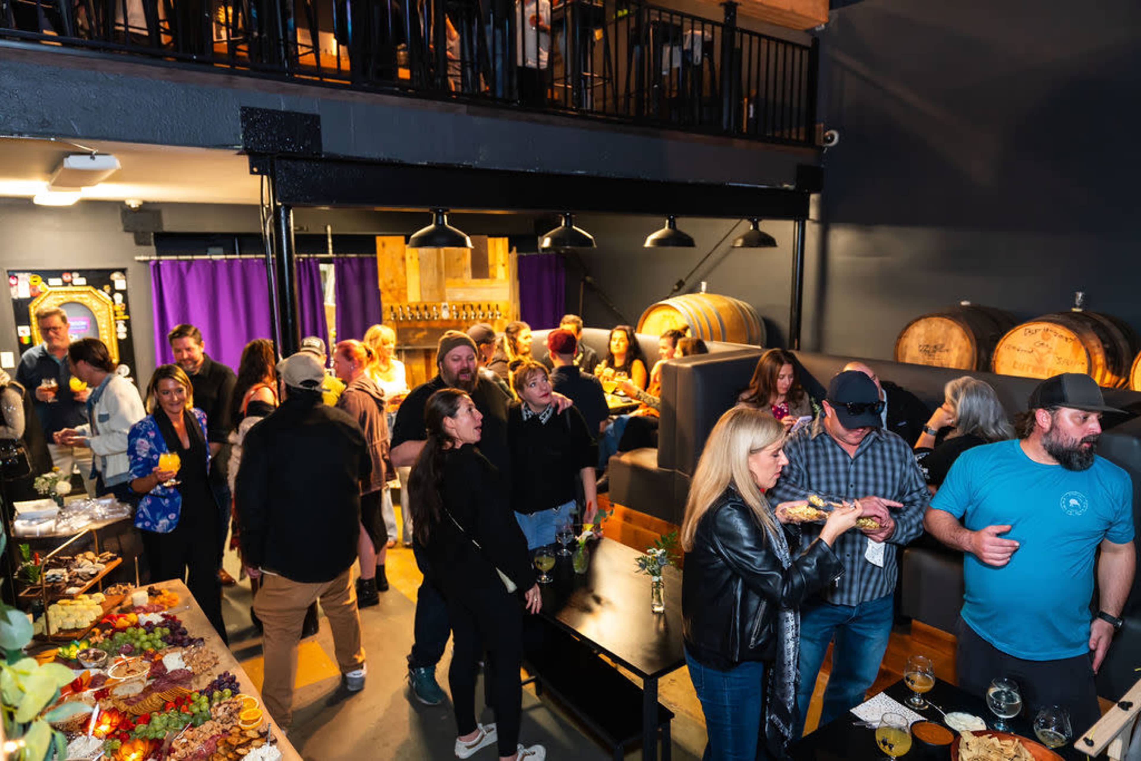 A crowd of people gathers in a lively bar setting, with tables of food and barrels in the background.