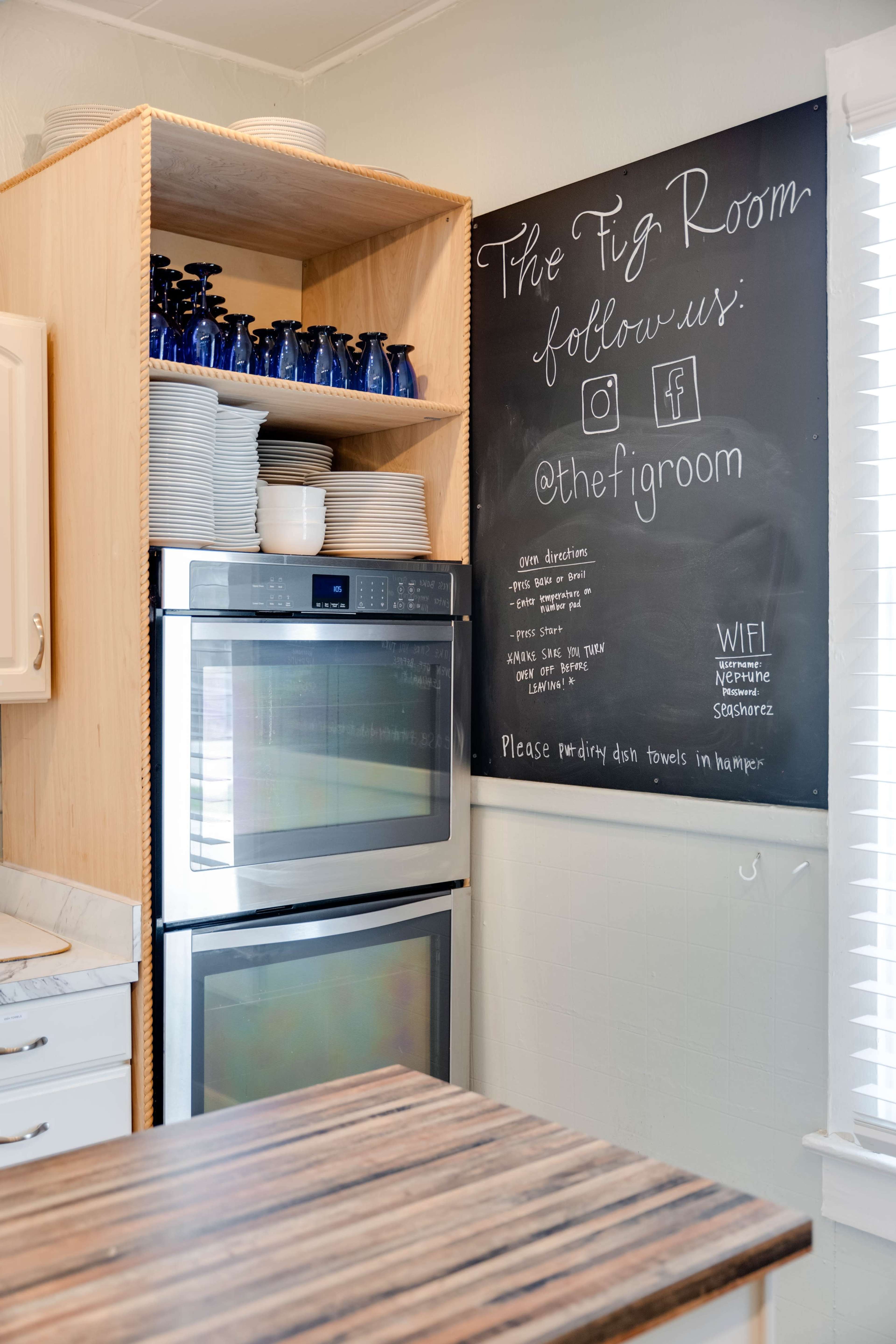A kitchen corner featuring a blackboard with social media information, shelves with blue glassware, and a stainless steel oven.